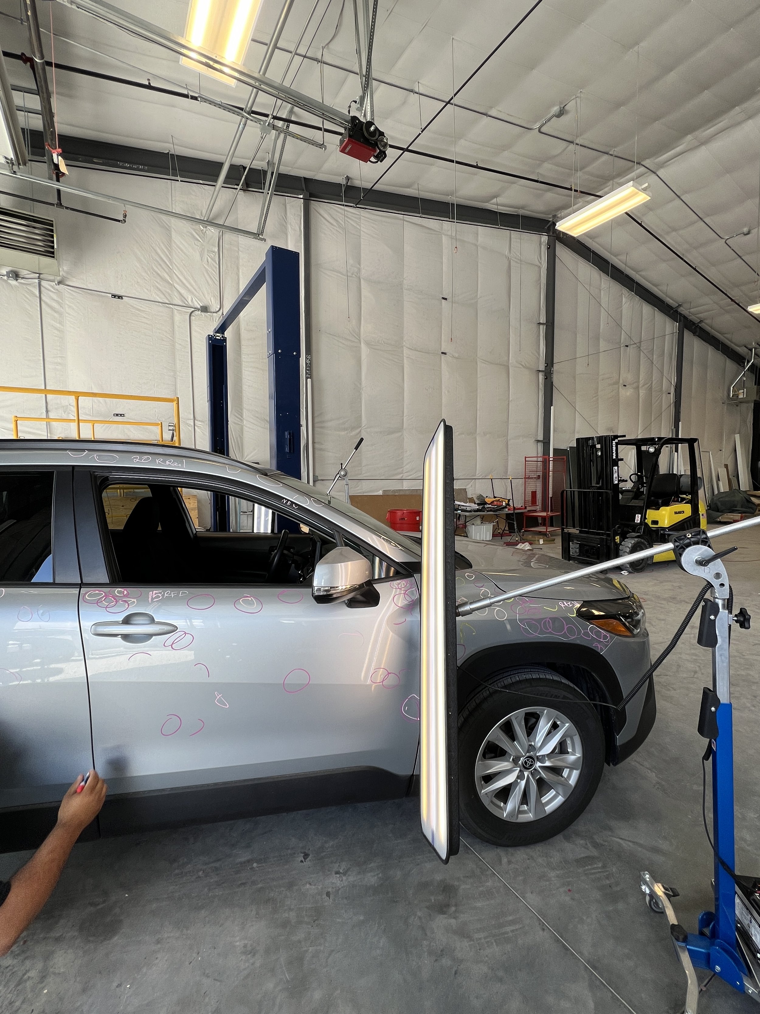 A silver Toyota car with pink and red markings being tested or repaired in an industrial garage, with a person’s hand visible on the left and equipment and tools in the background.