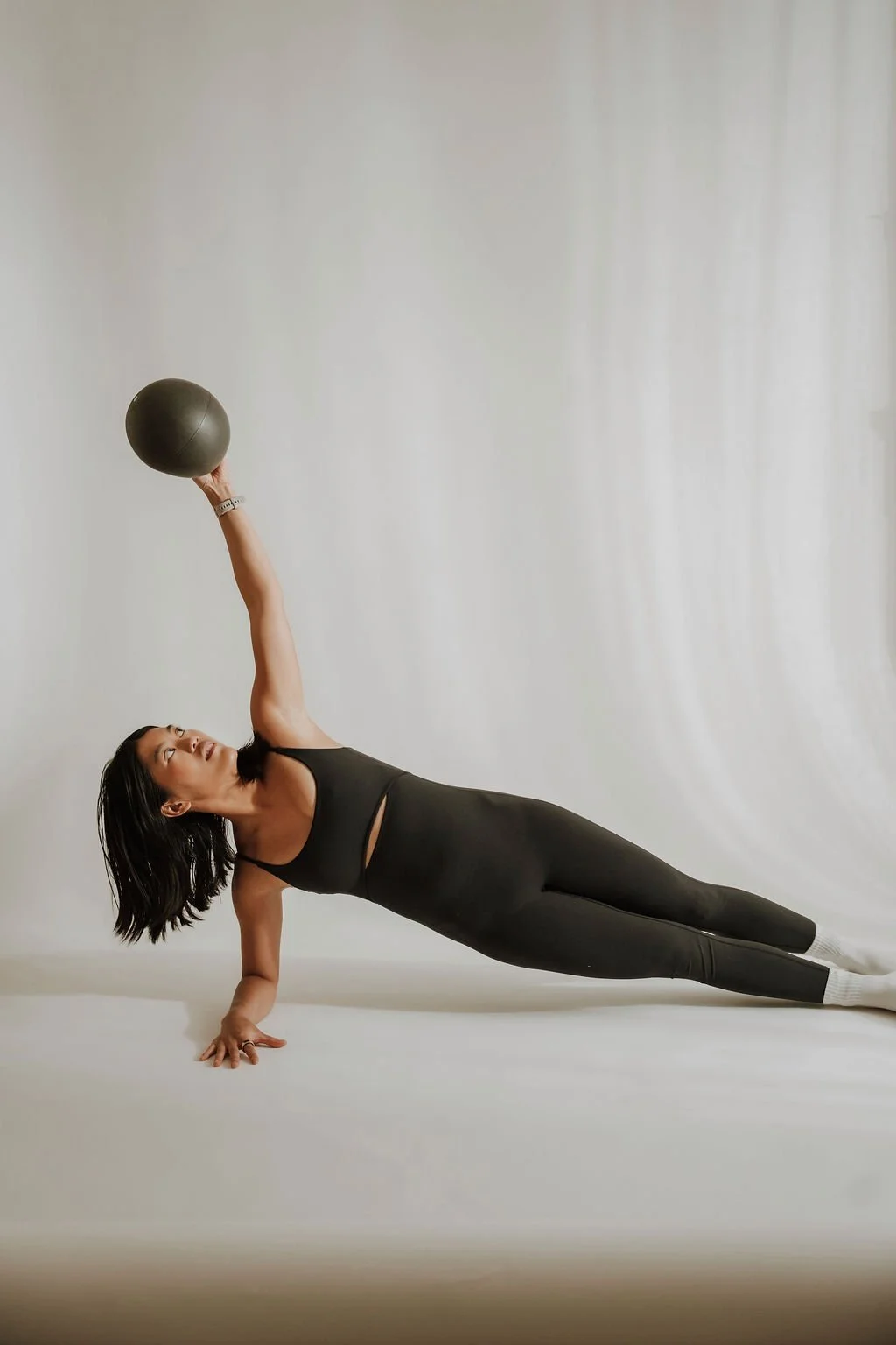 Woman performing a side plank with one arm raised, holding a black fitness ball, on a plain white background.