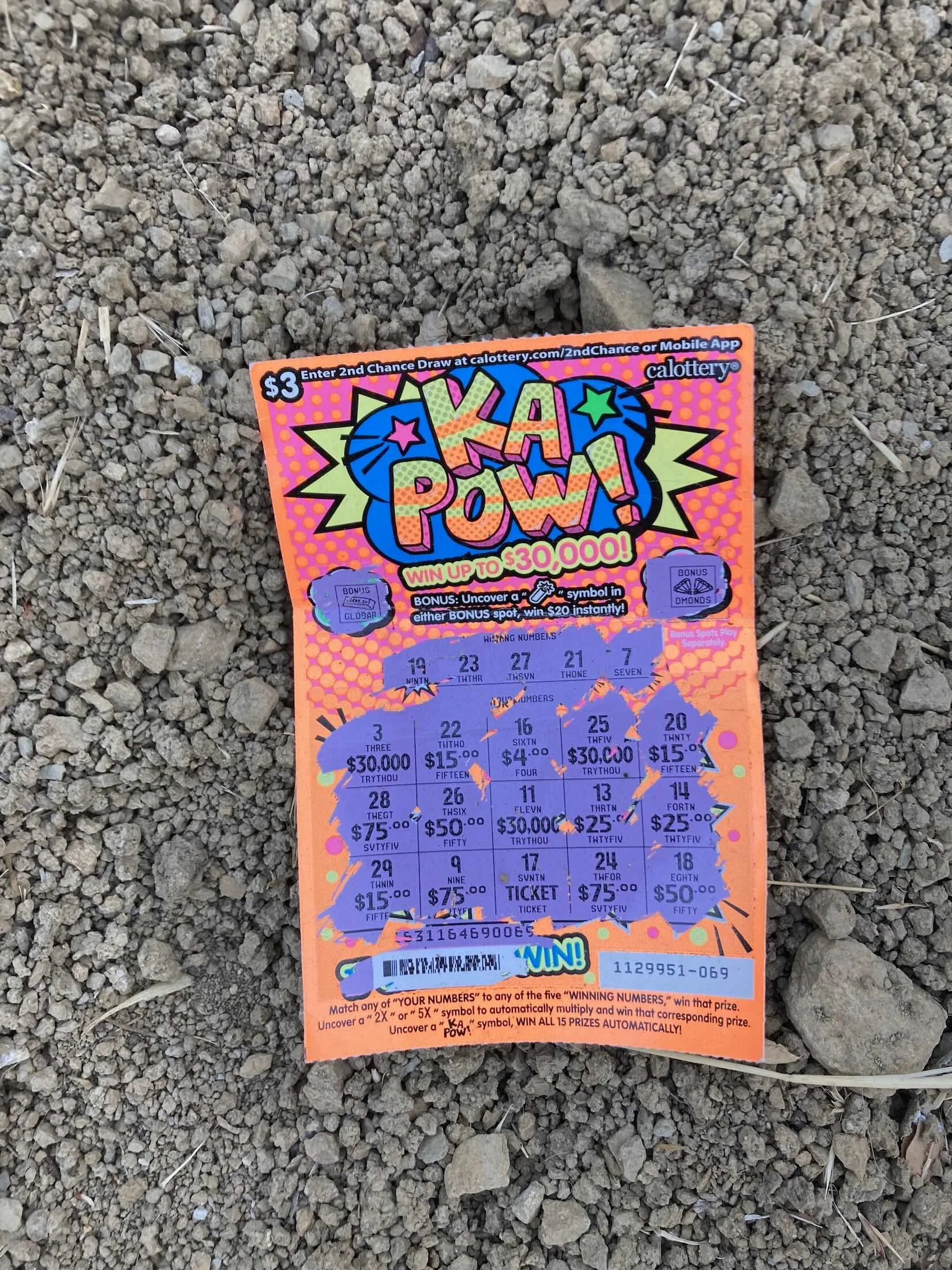 A bright orange scratch-off lottery ticket on the ground surrounded by dirt and small rocks. The ticket has the words 'KA POW!' in large, bold, block letters that appear to be exploding. The spaces have been scratched off.