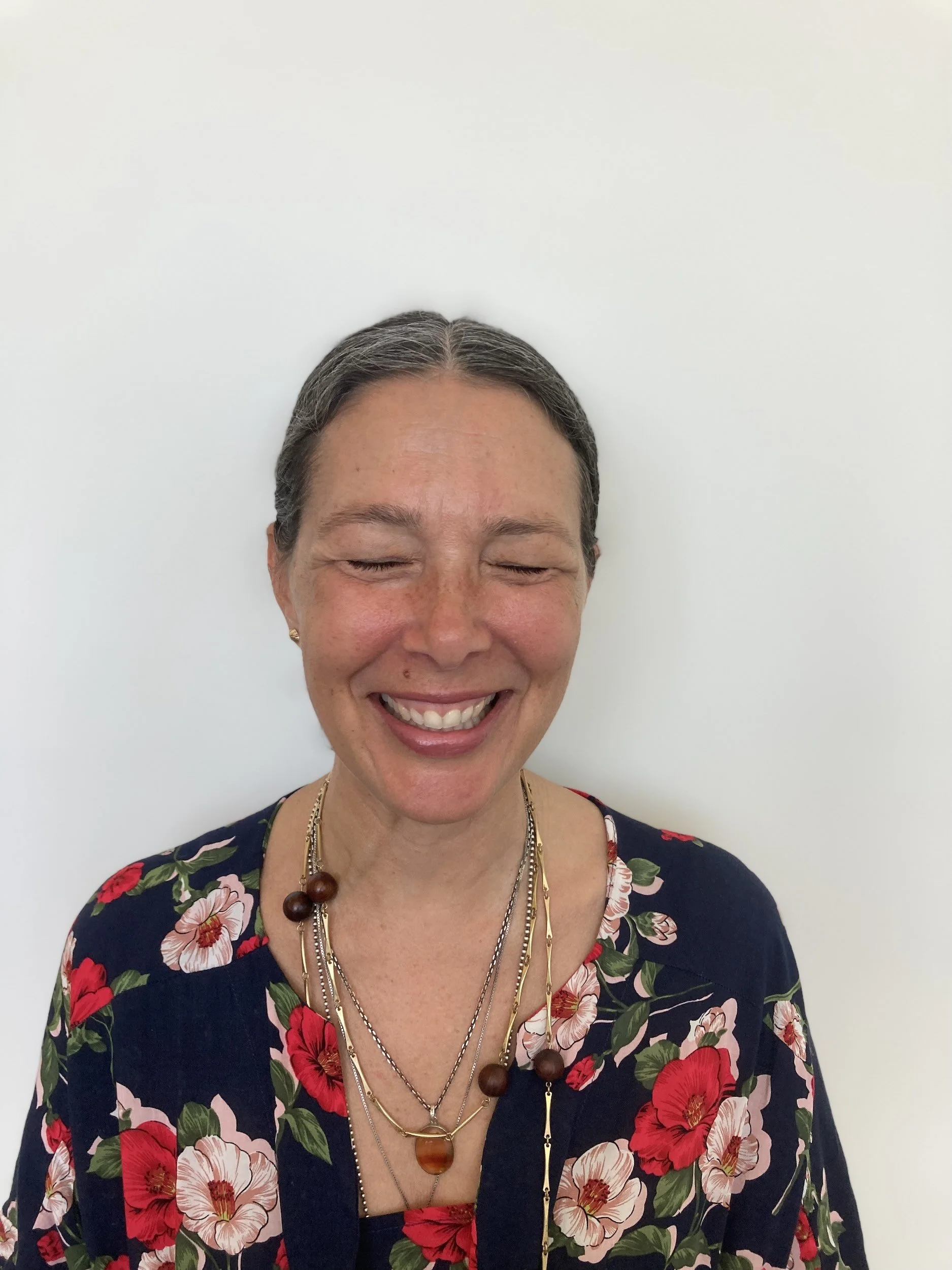 Author Megan Hirsch laughing with her eyes closed, wearing a floral dress and layered necklaces standing against a white wall.
