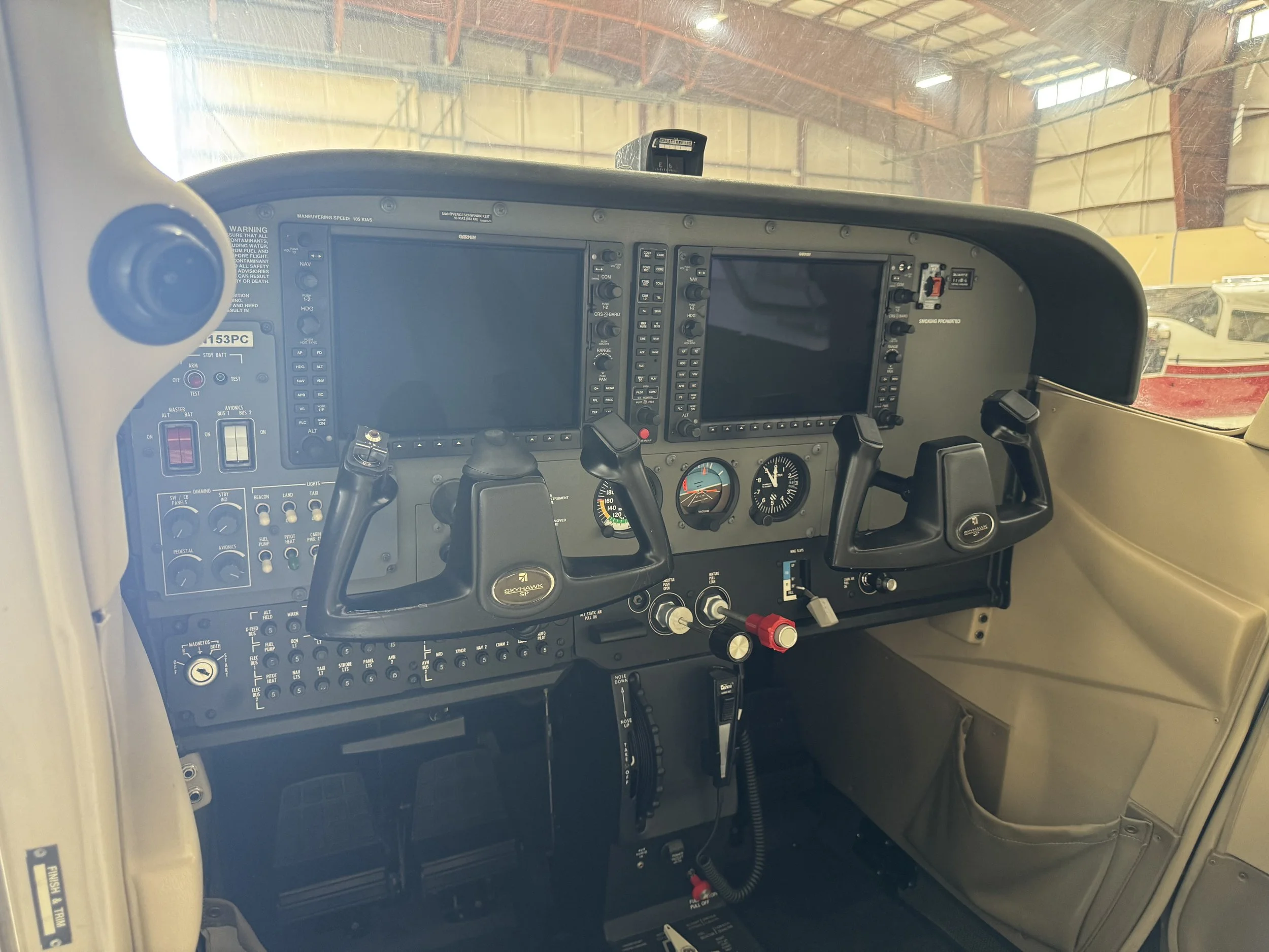 Interior cockpit of a light aircraft showing two large glass displays, various control switches, gauges, and a yoke with a Skyhawks logo.