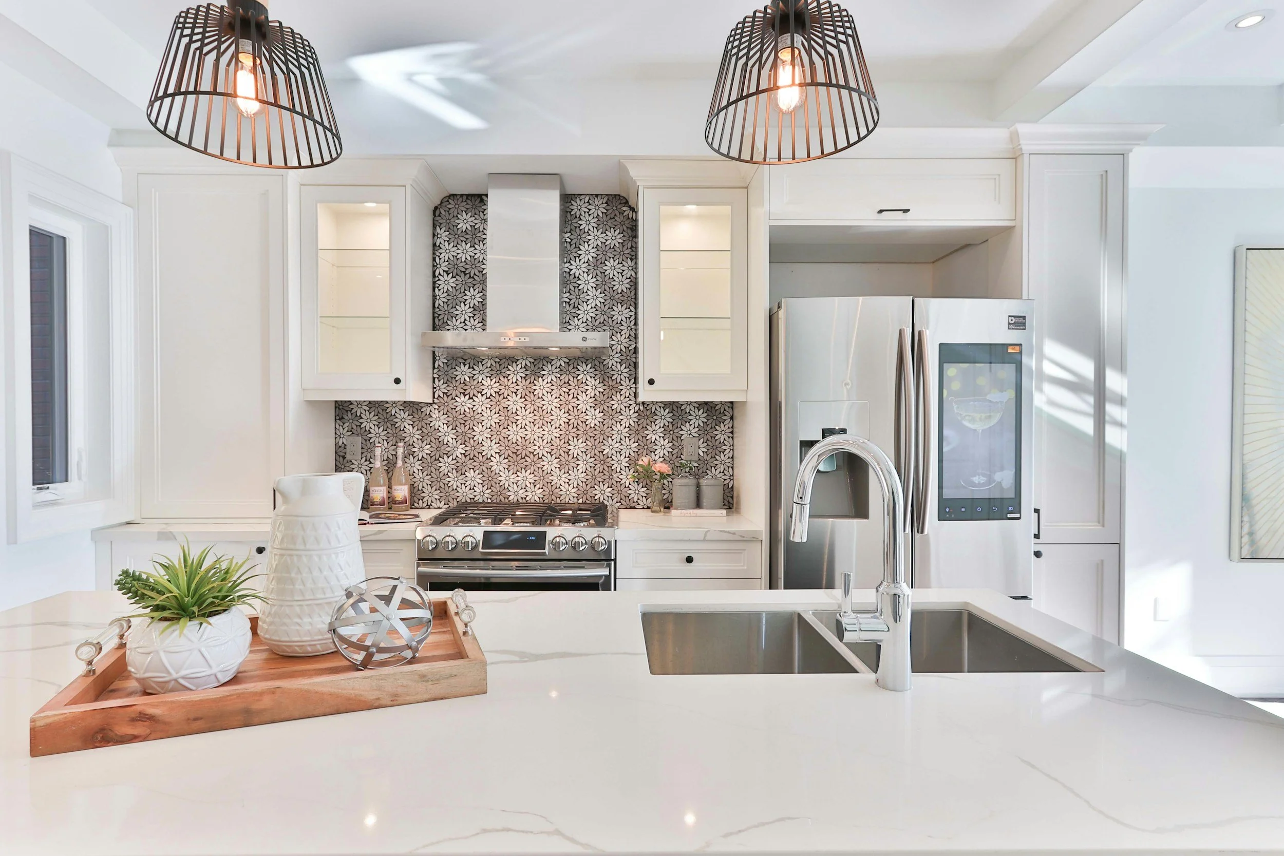 Modern white kitchen with black geometric pendant lights, stainless steel appliances, a patterned backsplash, and decorative plants on a white countertop