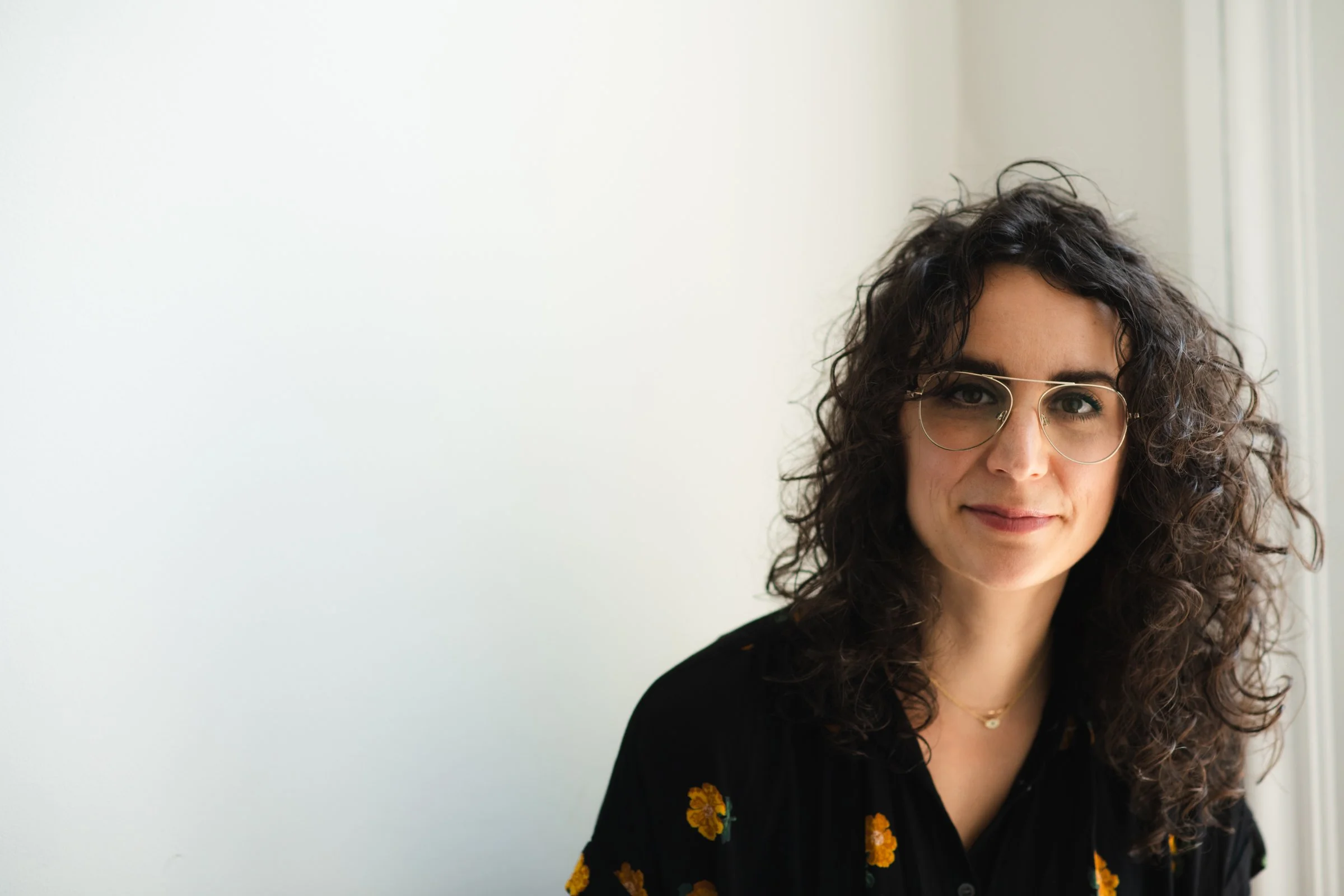A woman with curly dark hair wearing glasses and a black shirt with yellow floral patterns, smiling softly near a window with natural light.