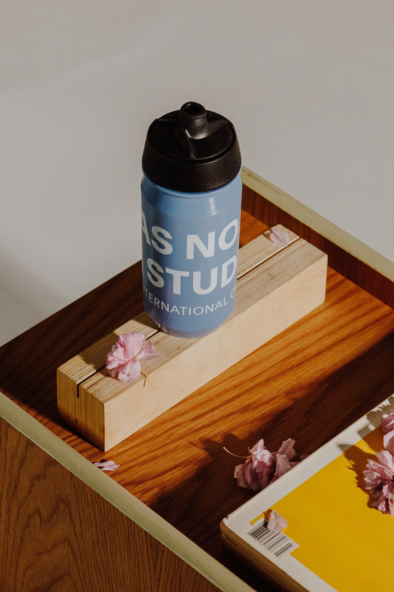 A blue water bottle with a black lid that has a spout, placed on a small wooden block on a wooden table. There are pink flower petals scattered on the table and a yellow notebook underneath a white tray.