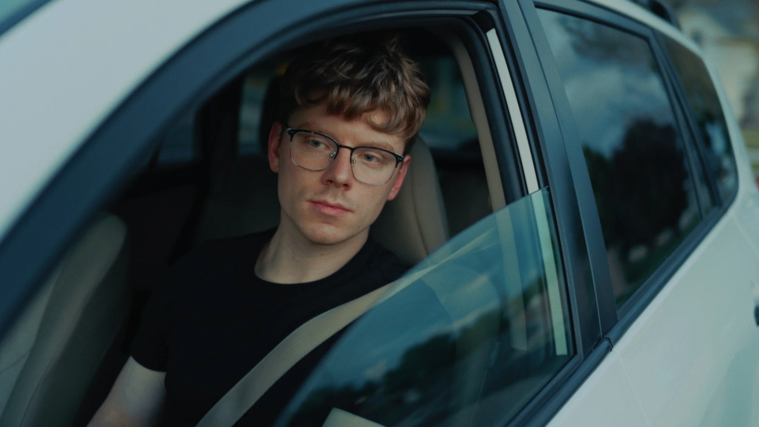 A young man with glasses and curly hair looking out of a car window.