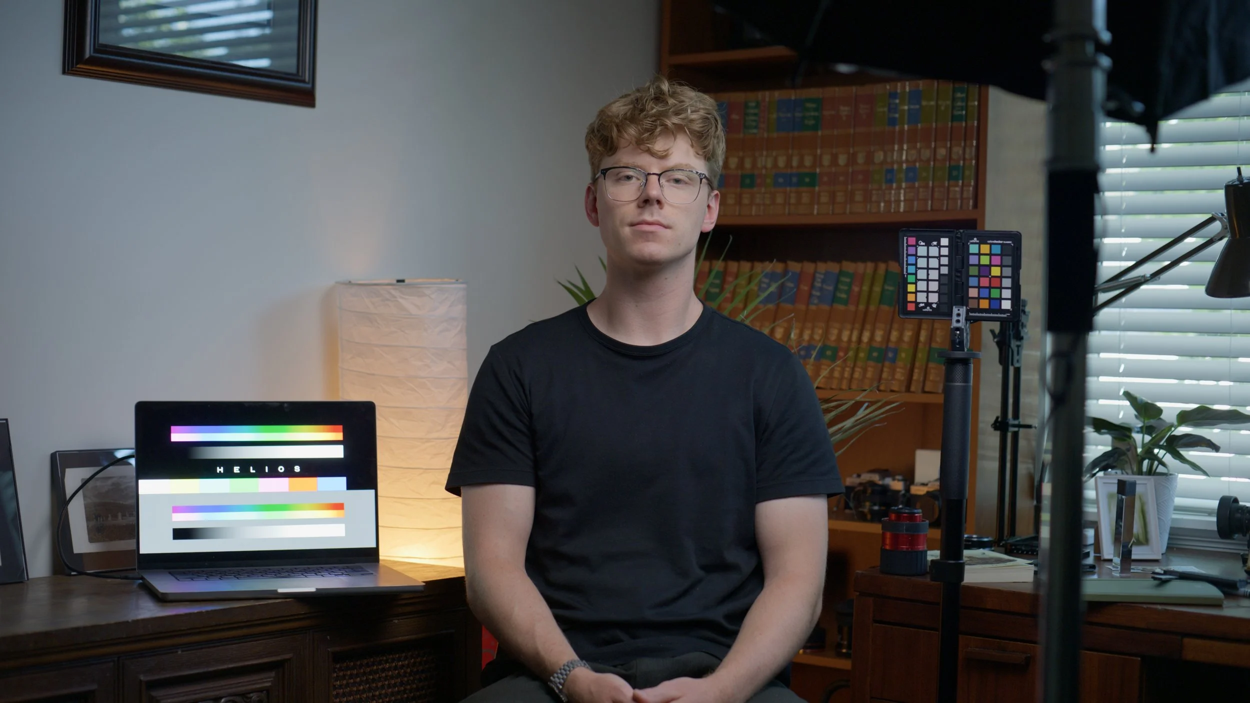 Young man with curly hair and glasses sitting at a desk in a home studio. Behind him are shelves with books, a laptop displaying a color test pattern, and photography equipment including a light diffuser, color checker, and camera mount. The room has natural light coming through blinds.