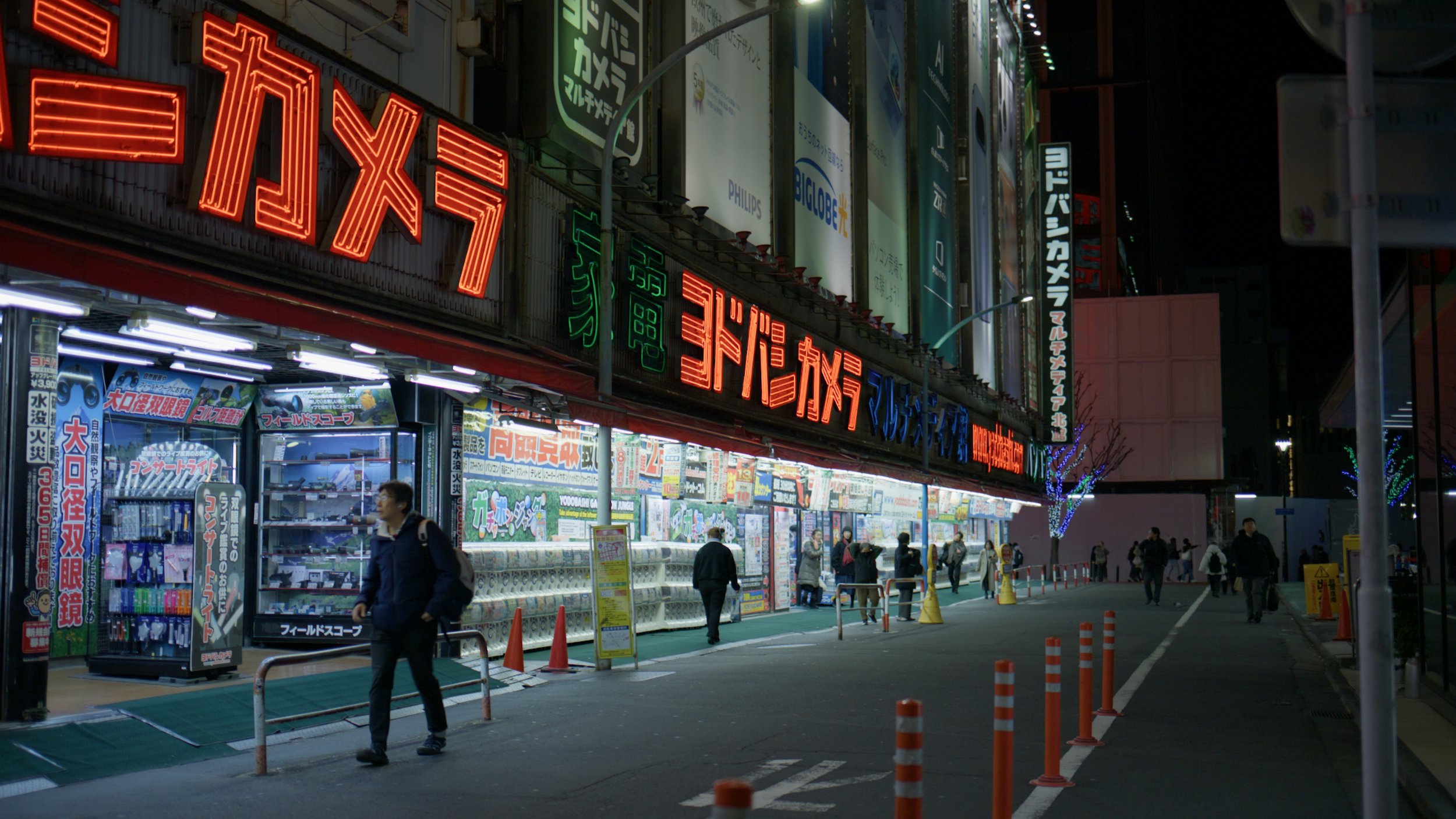 Night scene of a busy street with illuminated signs in Japanese, a storefront with advertisements, and people walking on the sidewalk.