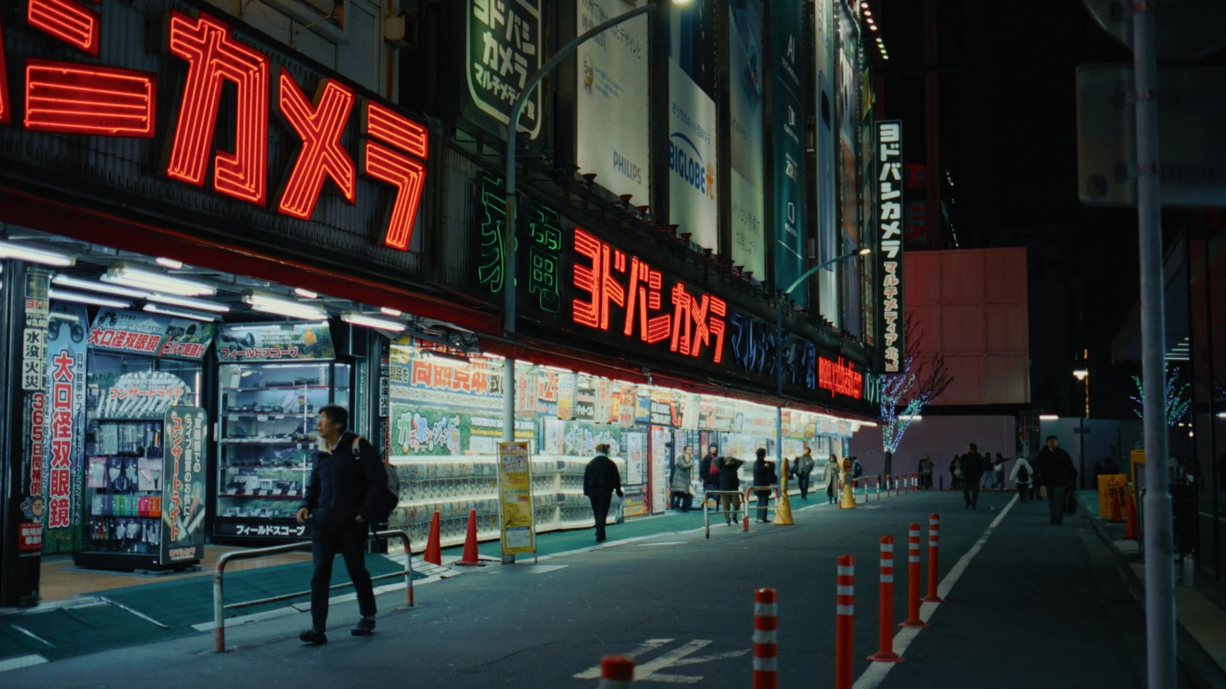 Nighttime street scene in Japan with illuminated signs in Japanese and storefront with magazines and posters, several pedestrians walking, some orange traffic cones, and trees decorated with lights.
