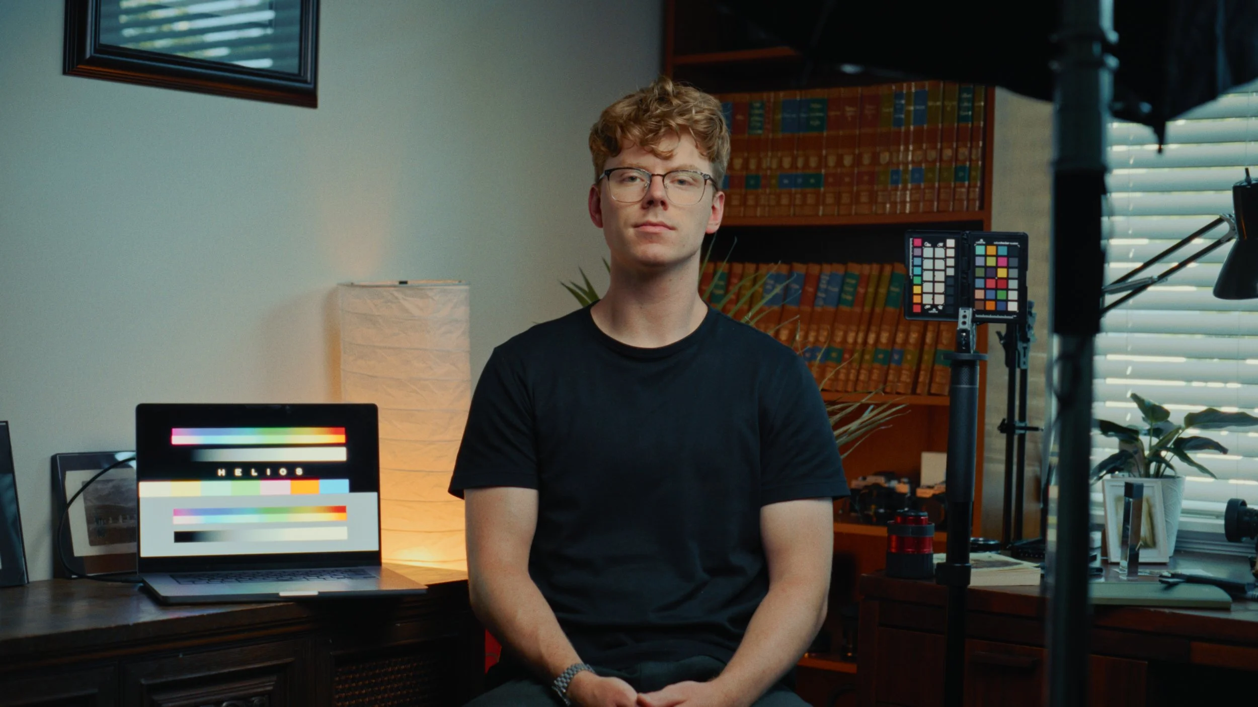 A young man with curly red hair and glasses sitting in a workspace surrounded by technical equipment, shelves full of books, a mirror, and a computer displaying a color test pattern.