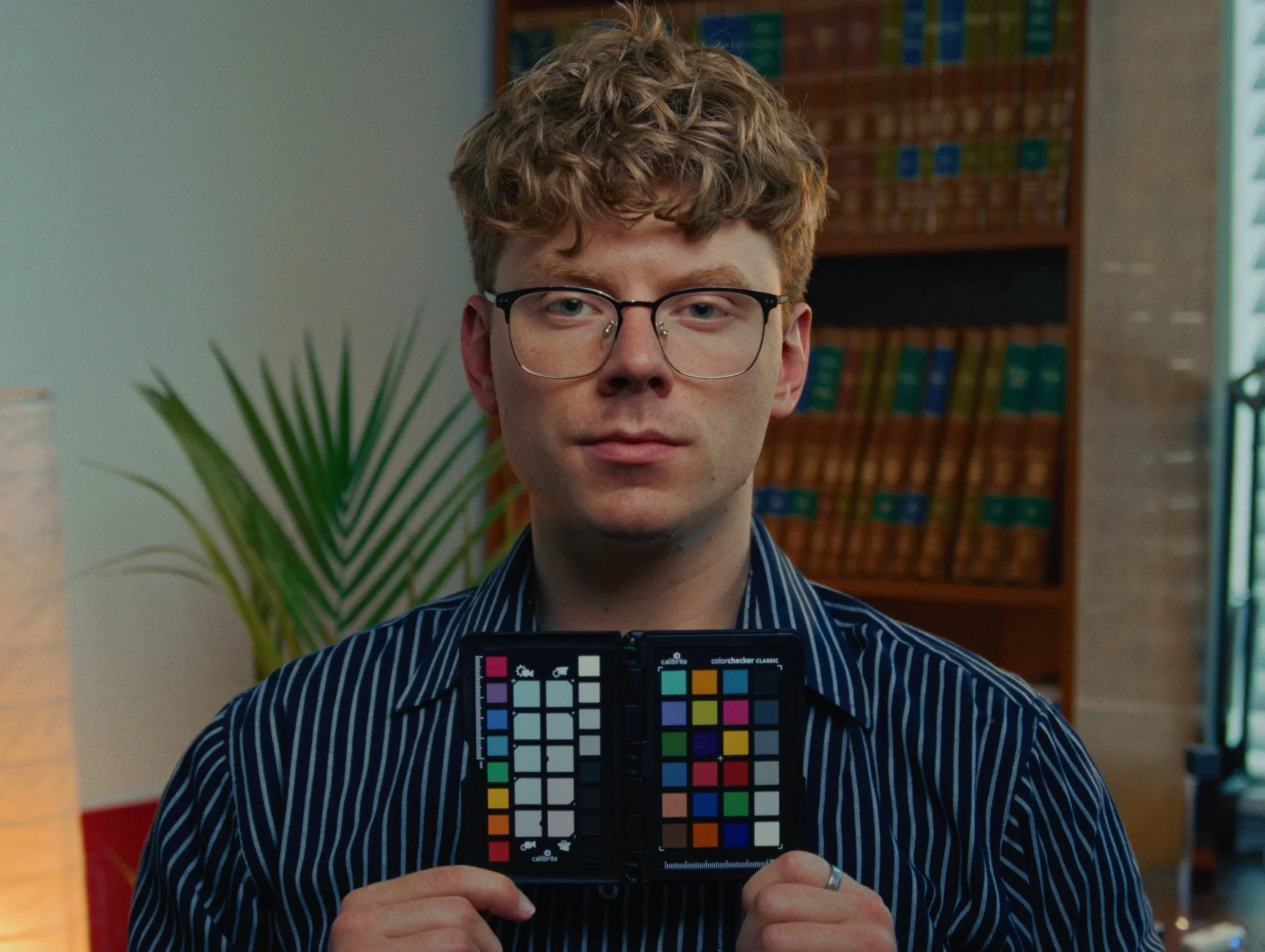 A man with curly red hair and glasses holding a color calibration chart in front of his face. He is wearing a striped shirt and is indoors, possibly in a library or office setting, with shelves of books in the background.