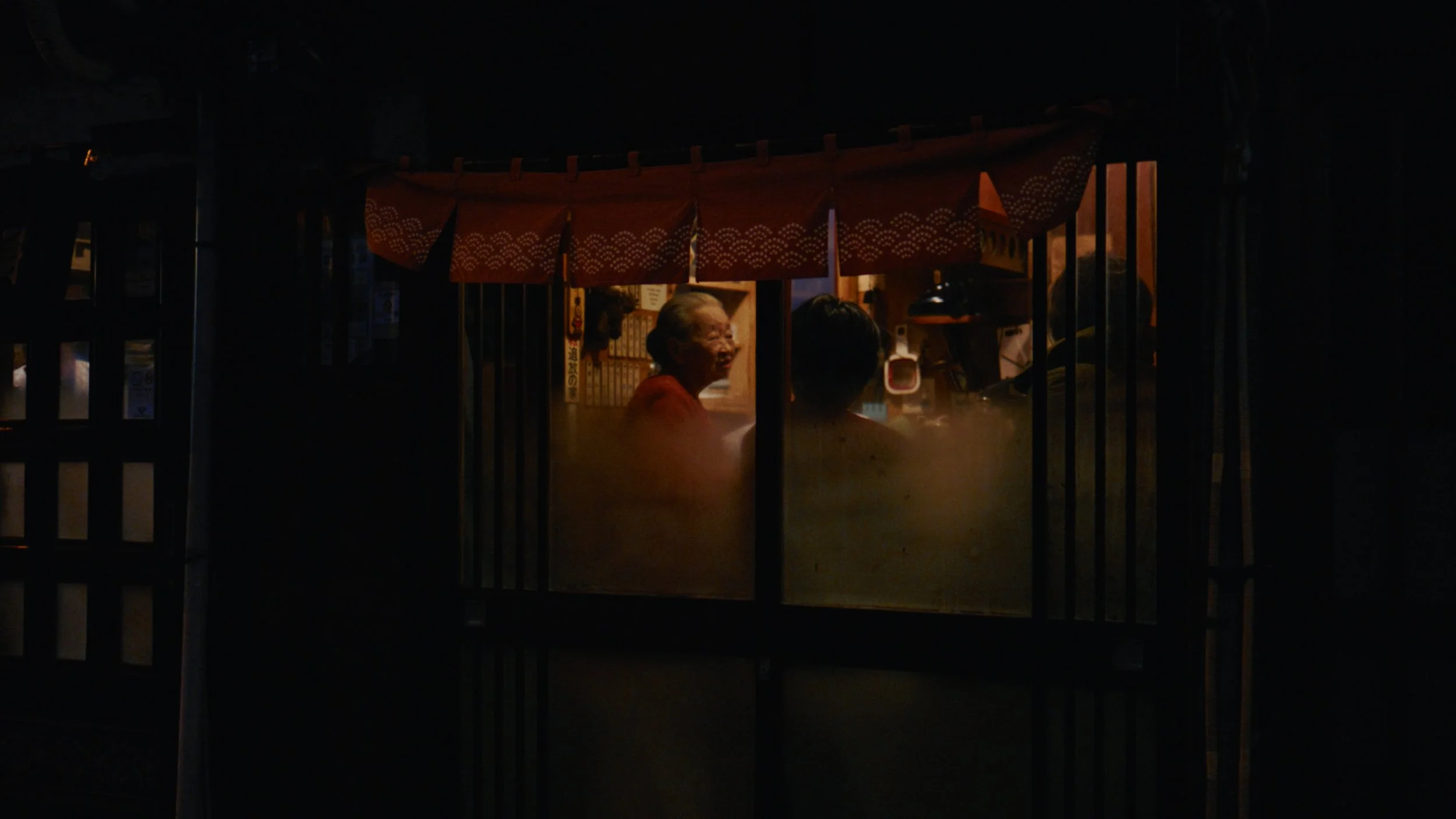 A nighttime scene of two people inside a small, illuminated Japanese-style booth, with one person smiling and the other person's back visible, framed by wooden slats and a red cloth with white patterns.