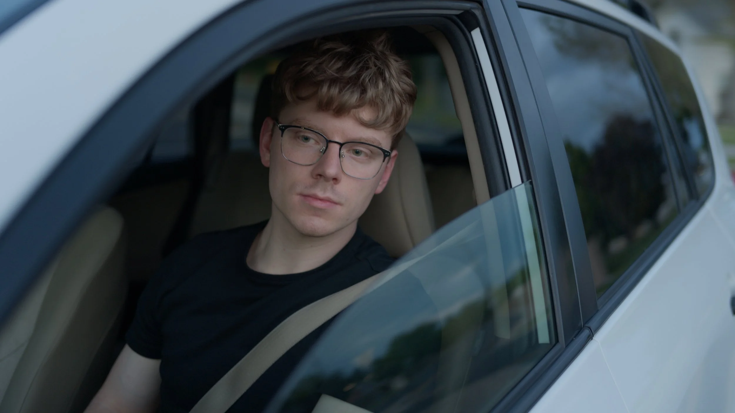 A young man with curly brown hair and glasses looking out the car window.