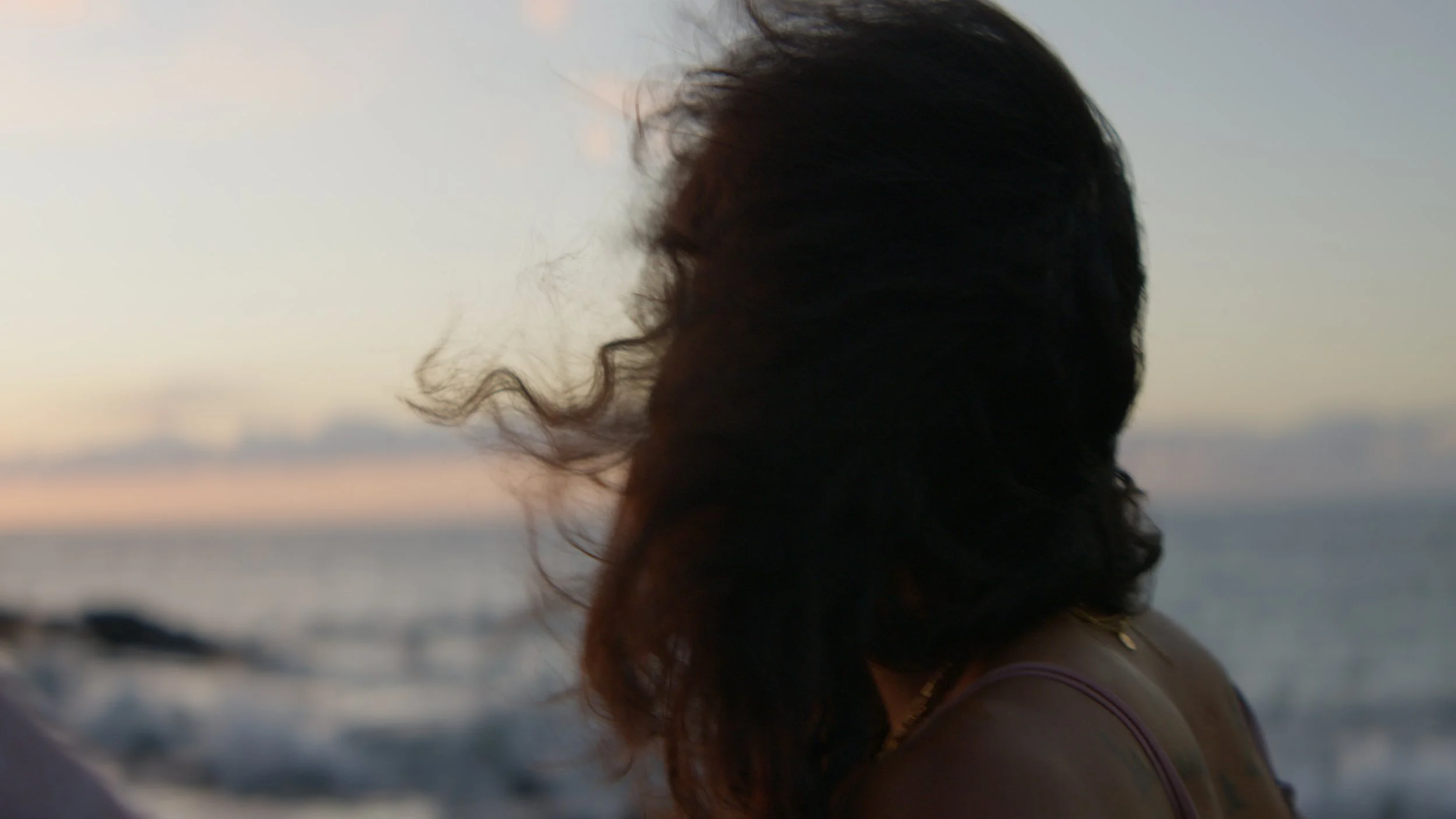A woman with curly hair standing near the water at sunset, facing sideways with her head slightly bowed.