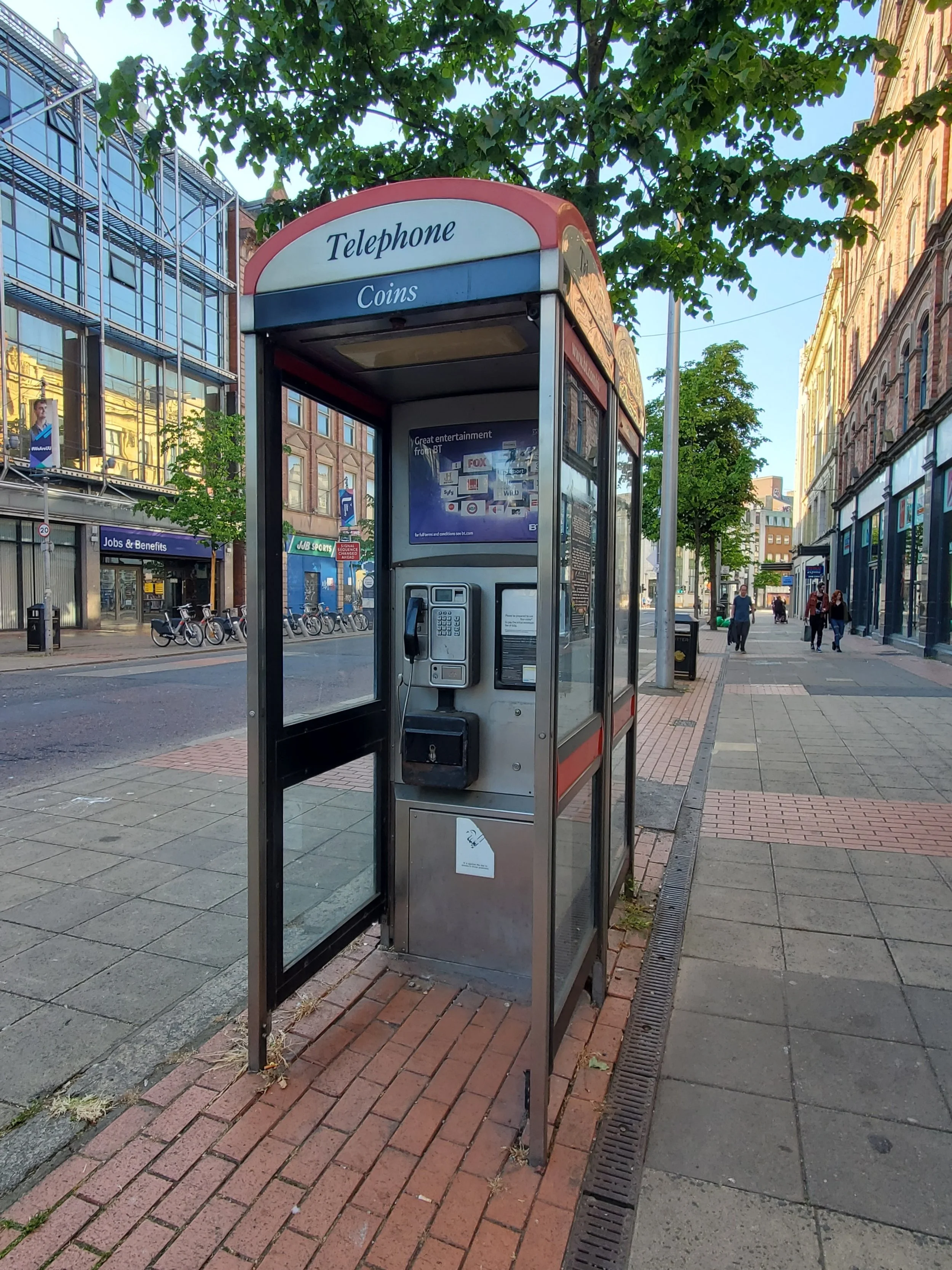 Old-fashioned telephone booth on a city sidewalk with a glass-covered phone inside; trees and buildings in the background.