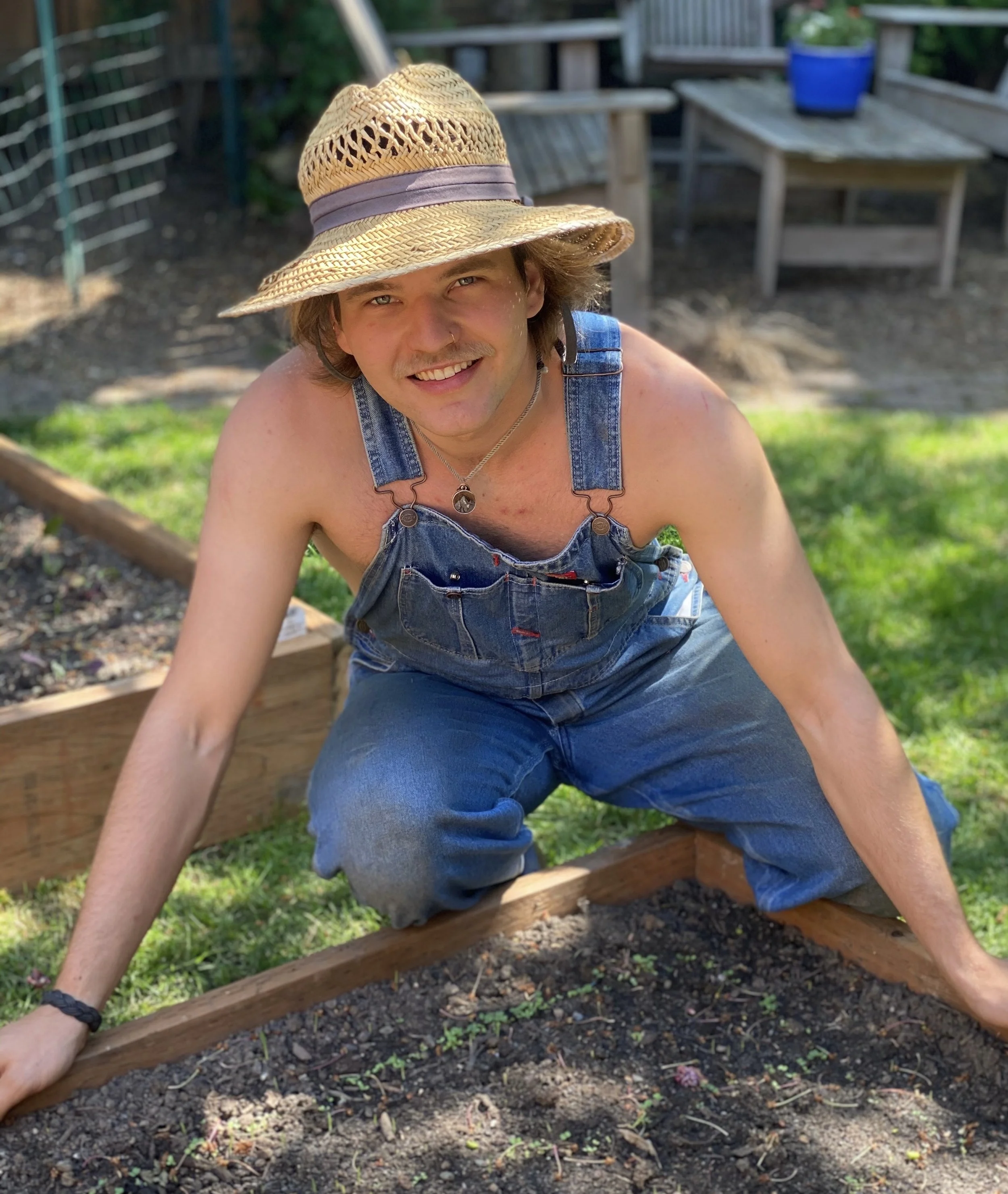 A young man wearing a straw hat, denim overalls, and a necklace, gardening outdoors in a sunny yard.