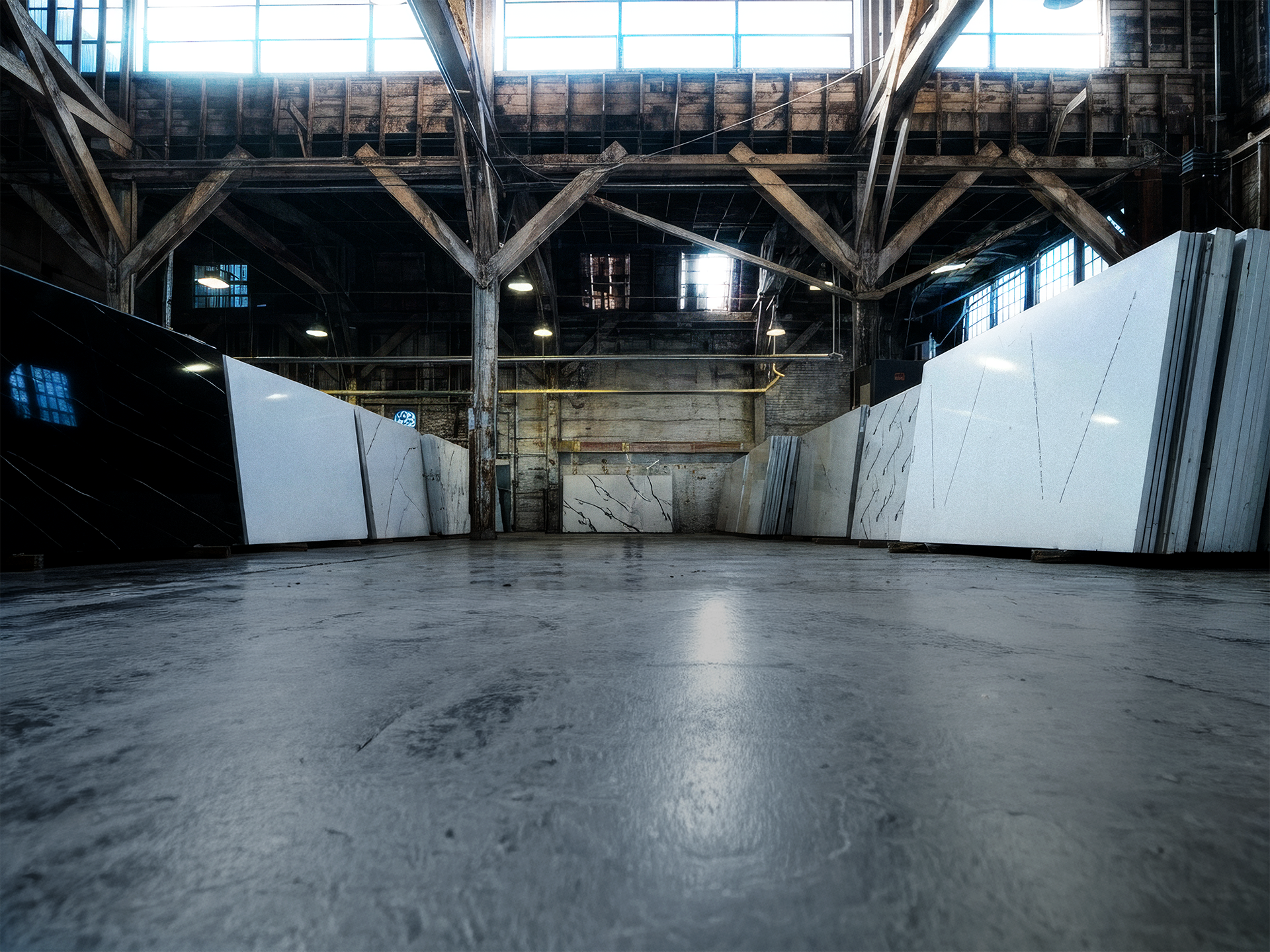 Interior view of a warehouse with large marble slabs leaning against the walls, wooden beams and industrial lighting overhead, and large windows at the top allowing natural light in.