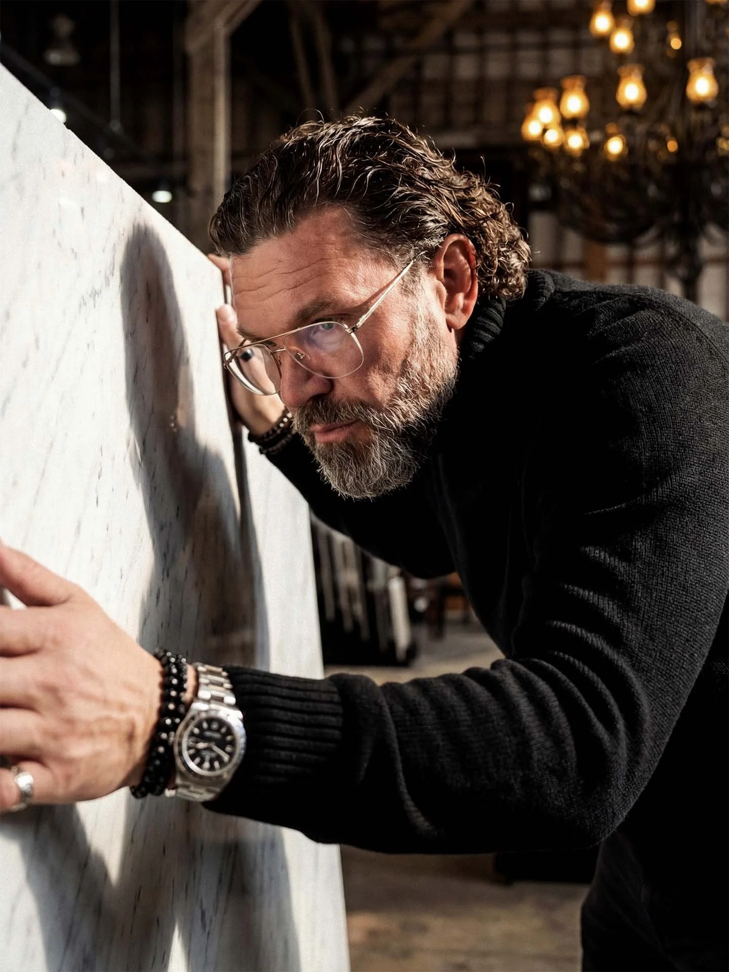 A man with glasses and a beard leaning close to a large light-colored textured surface, possibly a wall or canvas, with a background of warm hanging lights in an artist's studio or creative space.