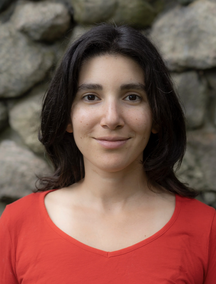 Close-up of a woman with shoulder-length dark hair wearing a red shirt, smiling softly, standing in front of a rocky background.