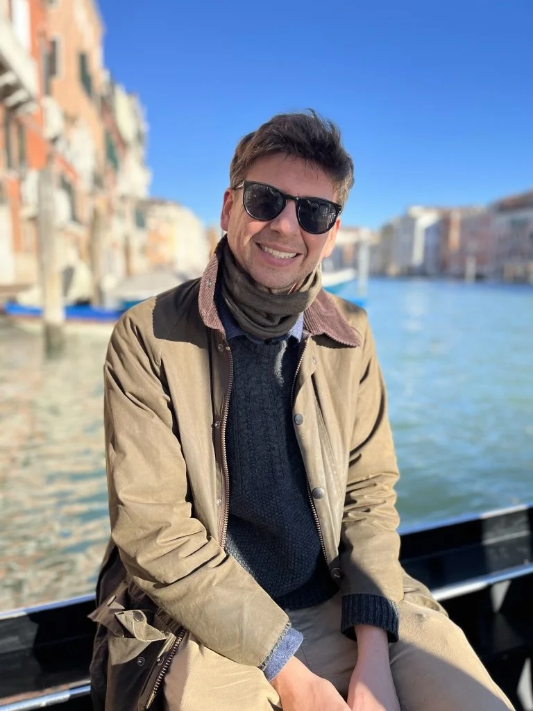 Smiling young man wearing sunglasses, a brown jacket, and a scarf, sitting on a boat with a canal and historical buildings in Venice, Italy, in the background on a sunny day.