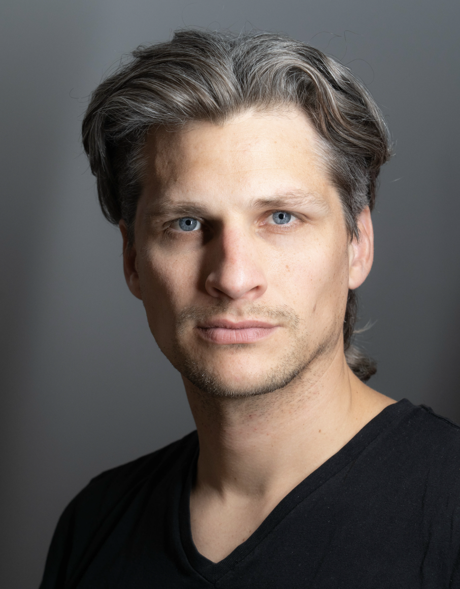 Close-up portrait of a young man with blue eyes, light skin, and dark blond hair, wearing a black shirt, against a gray background.