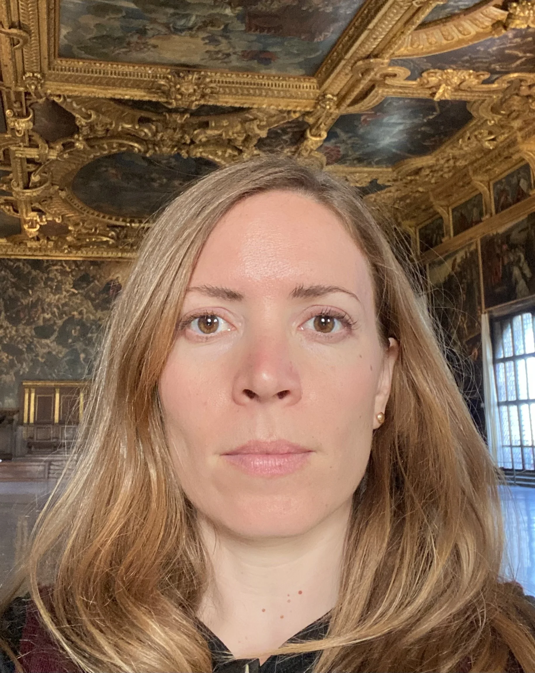 A woman with long, wavy, light brown hair looking directly at the camera, inside a grand room with ornate gold decorations and painted ceiling murals.