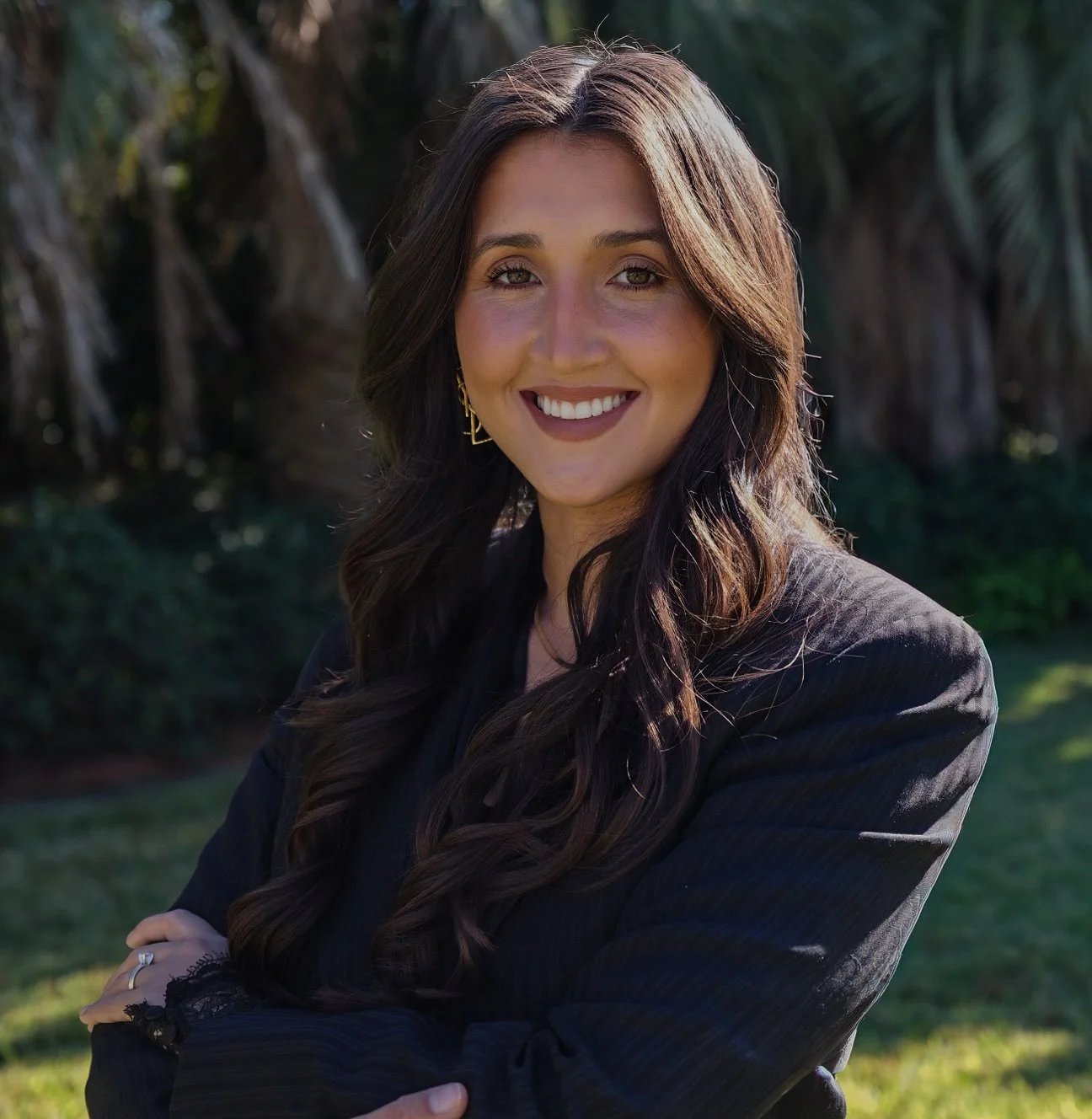 A woman with long dark wavy hair smiling outdoors in a garden with green bushes and trees.