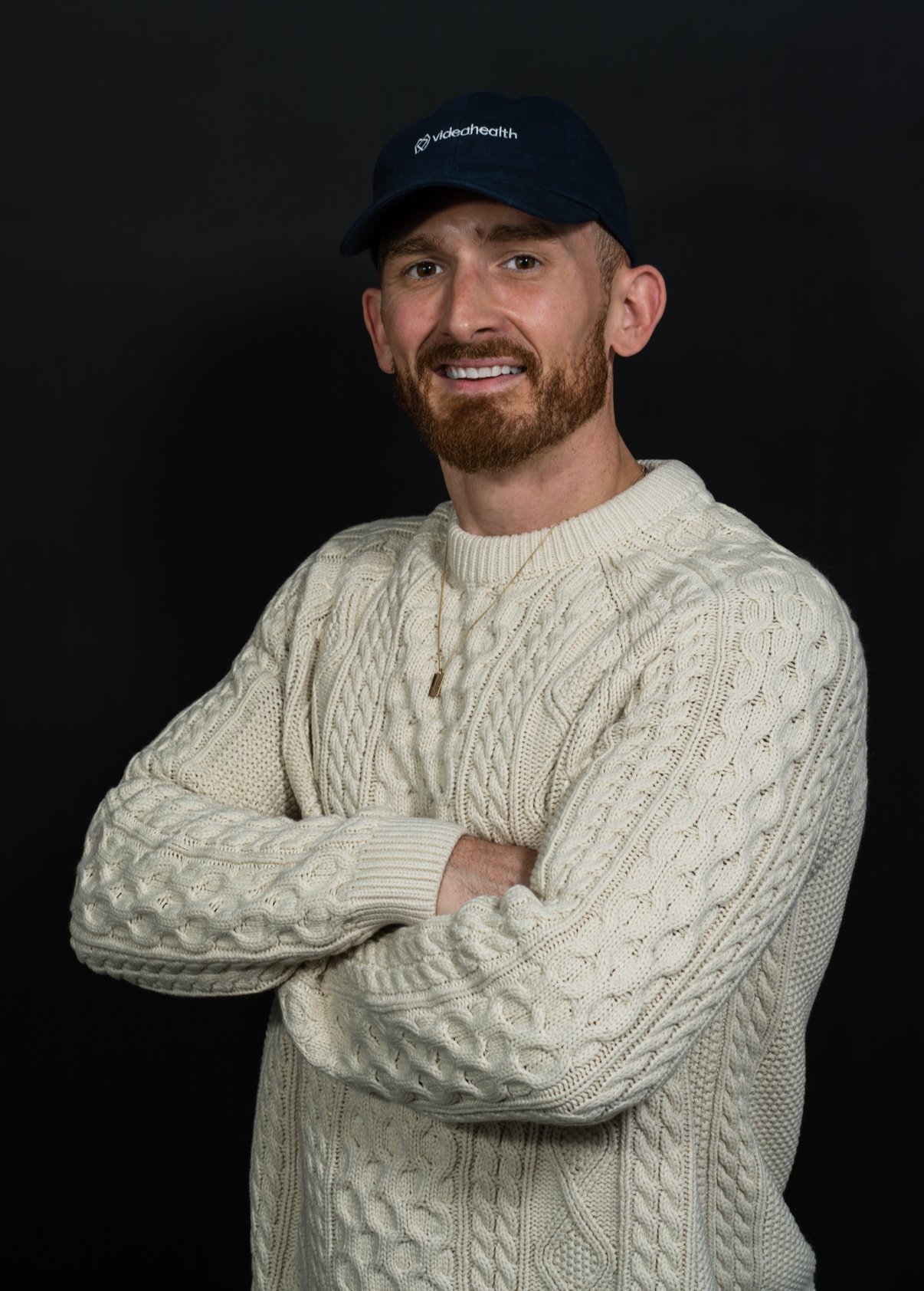 A man with a beard and mustache wearing a cream cable-knit sweater, a black baseball cap with 'video health' written on it, and a gold chain with a small pendant, standing against a black background with arms crossed and smiling.