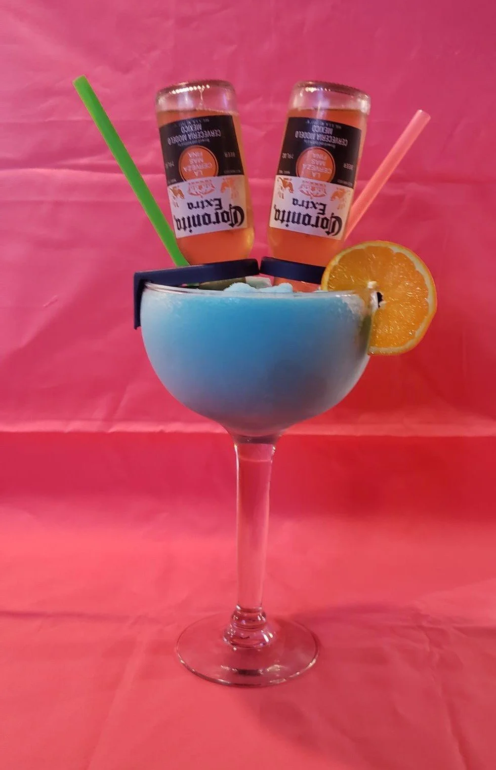 A colorful cocktail in a stemmed glass garnished with a slice of orange, with two miniature bottles of Corona beer and two straws against a pink background.