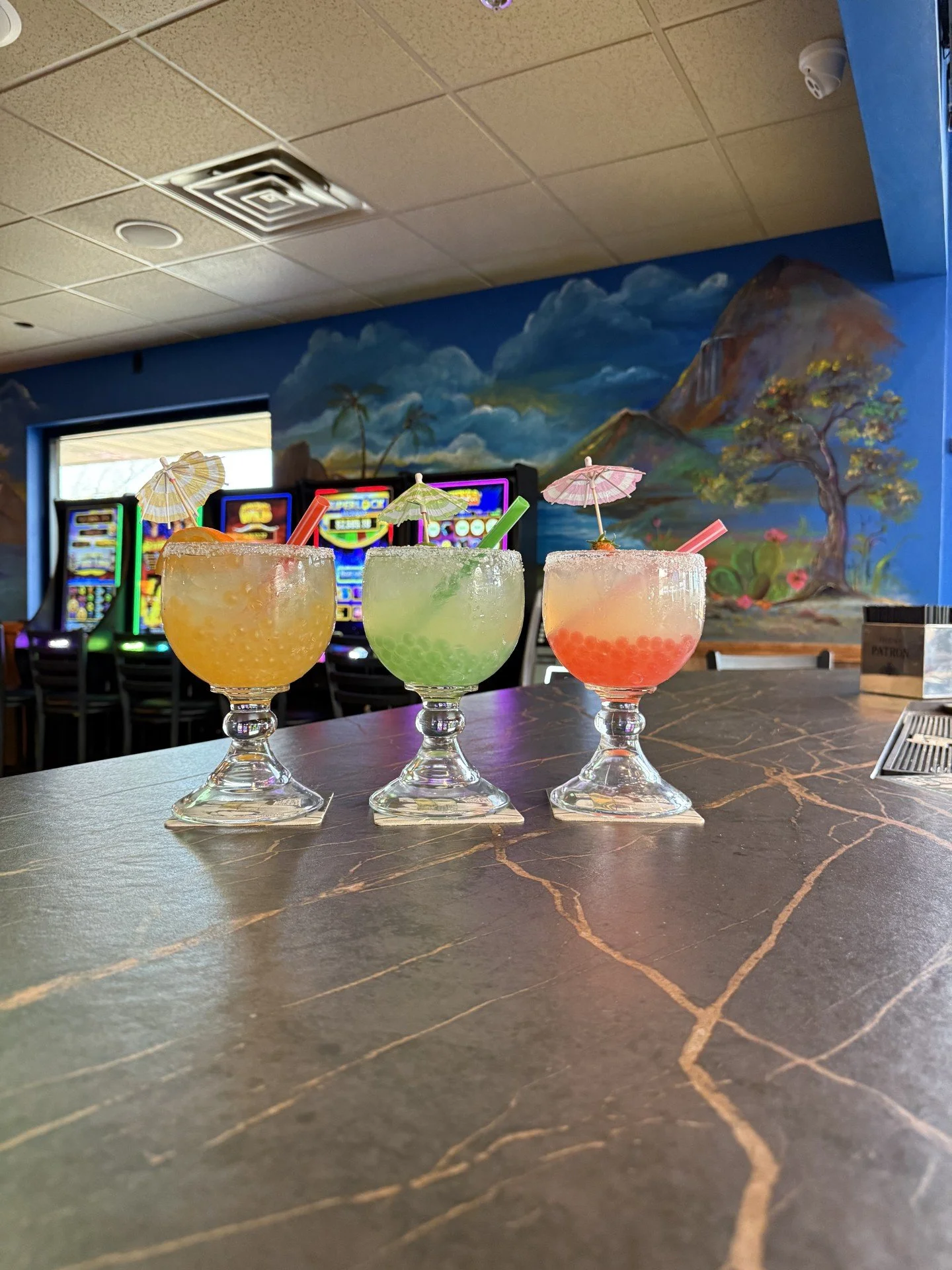 Three colorful cocktails with umbrellas and straws on a bar counter, with a mural of a tropical landscape and slot machines in the background.