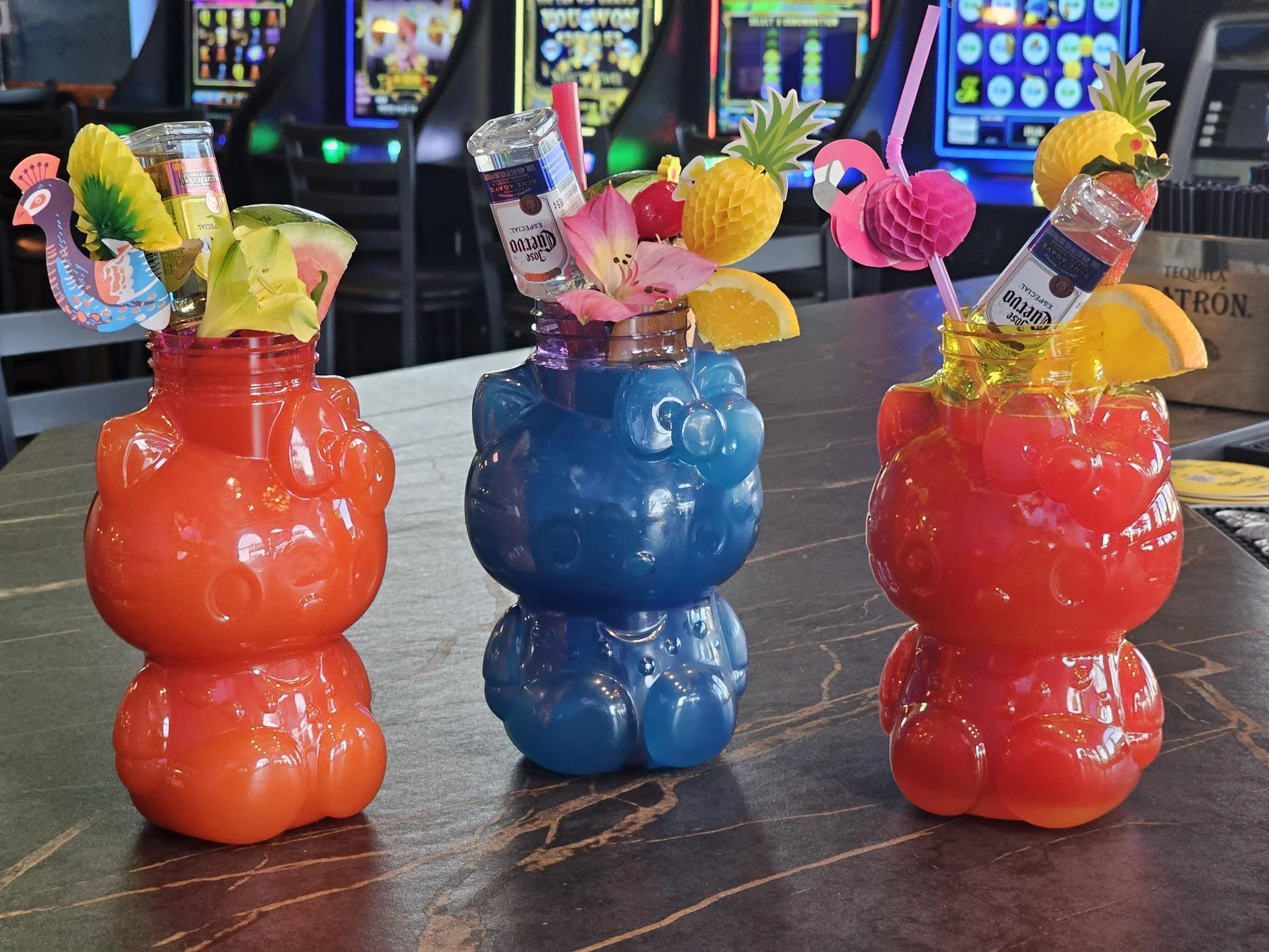 Three bear-shaped tiki glasses filled with colorful cocktails, garnished with fruit and small decorative items, on a dark countertop in front of slot machines.