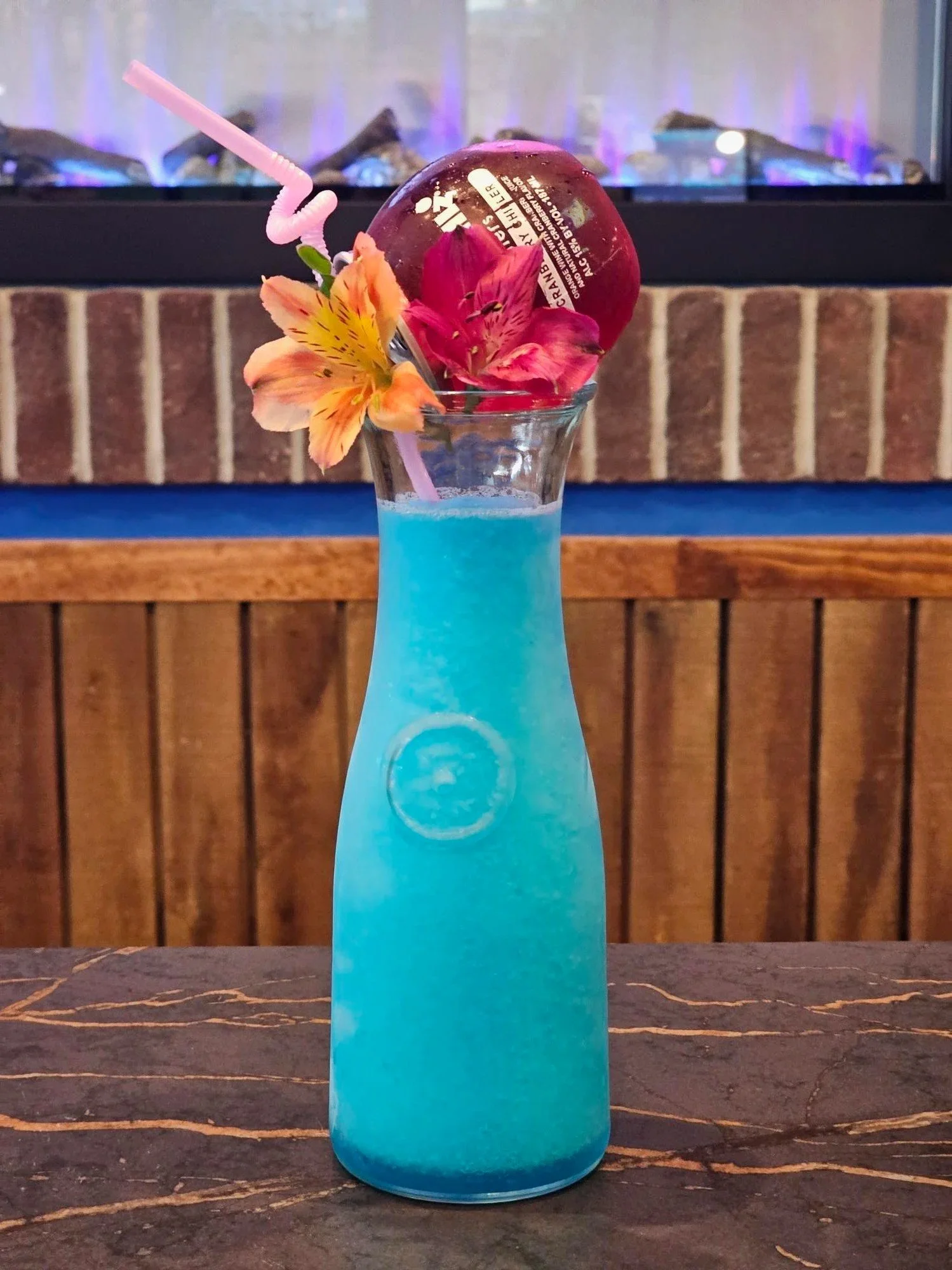 Blue cocktail in a clear beach-shaped glass with a flower garnish, a pink straw, and a cherry on top, placed on a dark marble table with a brick fireplace in the background.