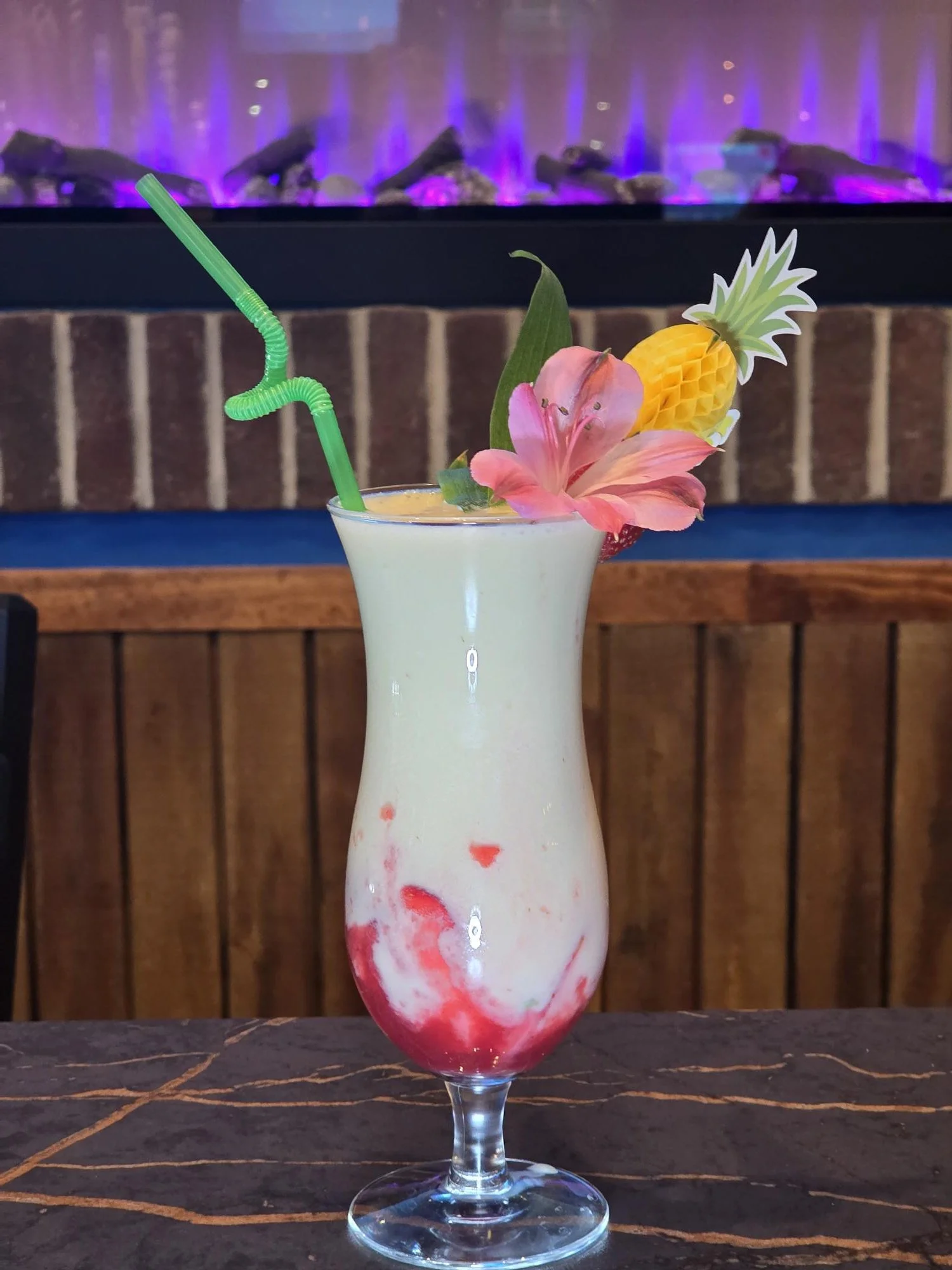 A tropical cocktail with a green bendy straw, garnished with a pink lily, a paper pineapple, and a pink flower, served in a tall, curvy glass on a dark marble surface.