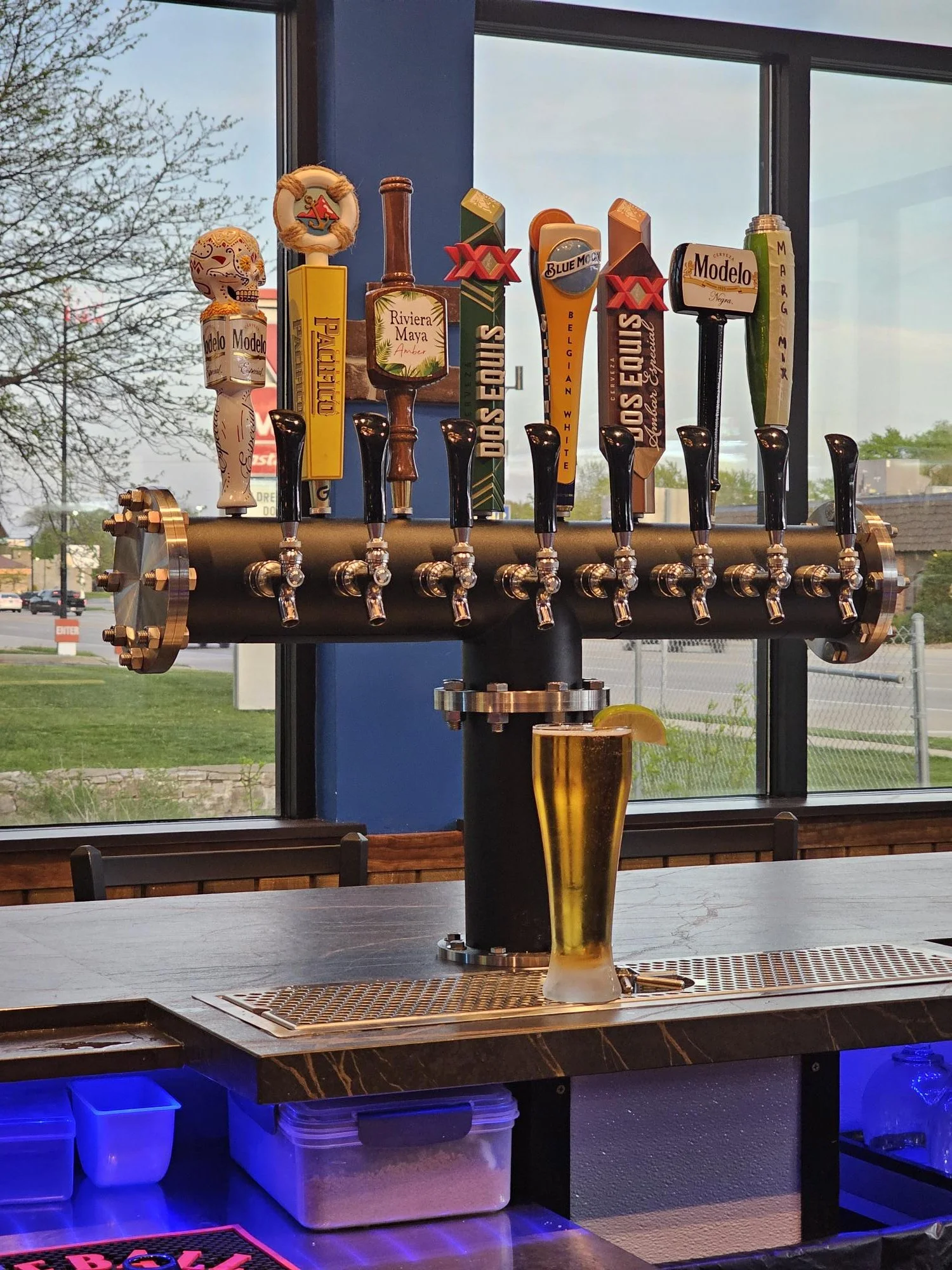 A beer tap display with multiple beer handles on a black and brass tap system behind a bar. In front, there is a glass of beer with a lemon wedge on the rim.