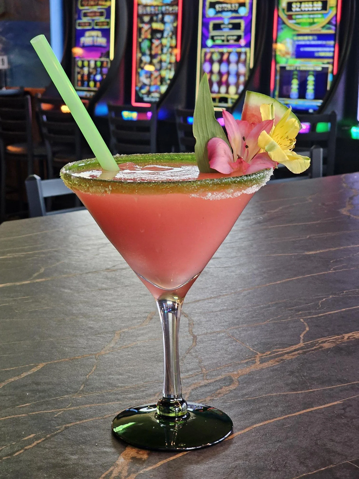 A pink cocktail in a martini glass garnished with pink and yellow flowers, a green leaf, and a green straw, set on a dark wood table with slot machines in the background.