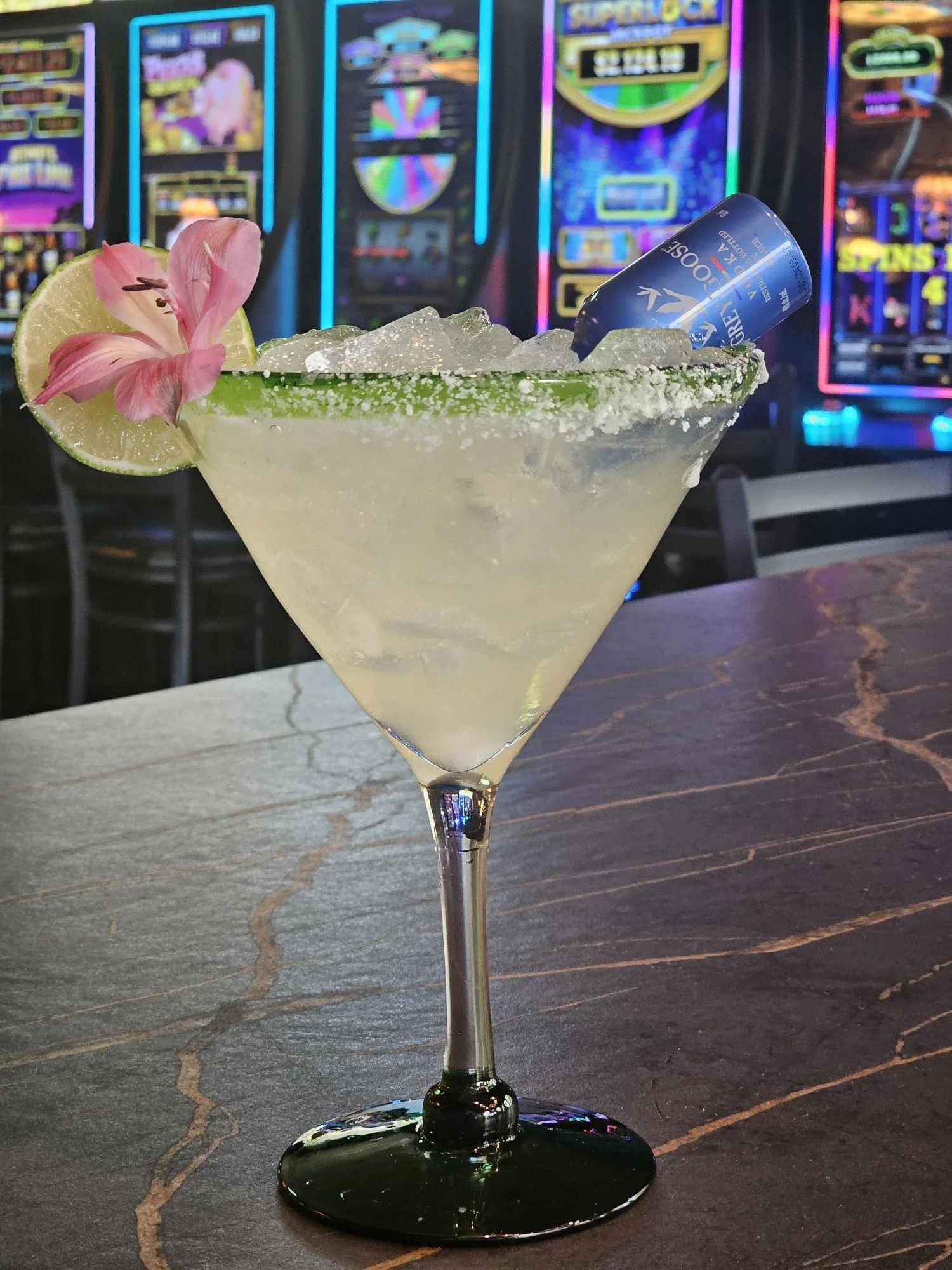 A cocktail glass filled with a light-colored drink and ice cubes, garnished with a lime wedge and a pink flower, with a small blue can of Coors Light beer inside the glass base. The background features a casino with colorful slot machines.