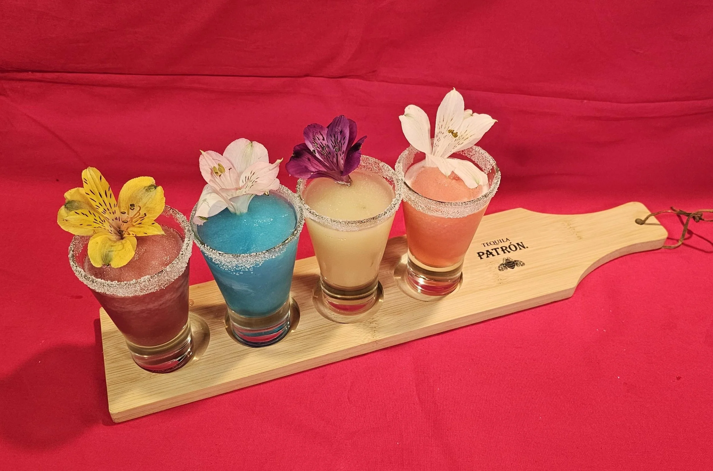 Four colorful cocktails with sugar-rimmed glasses and edible flowers on a wooden paddle, against a pink background.