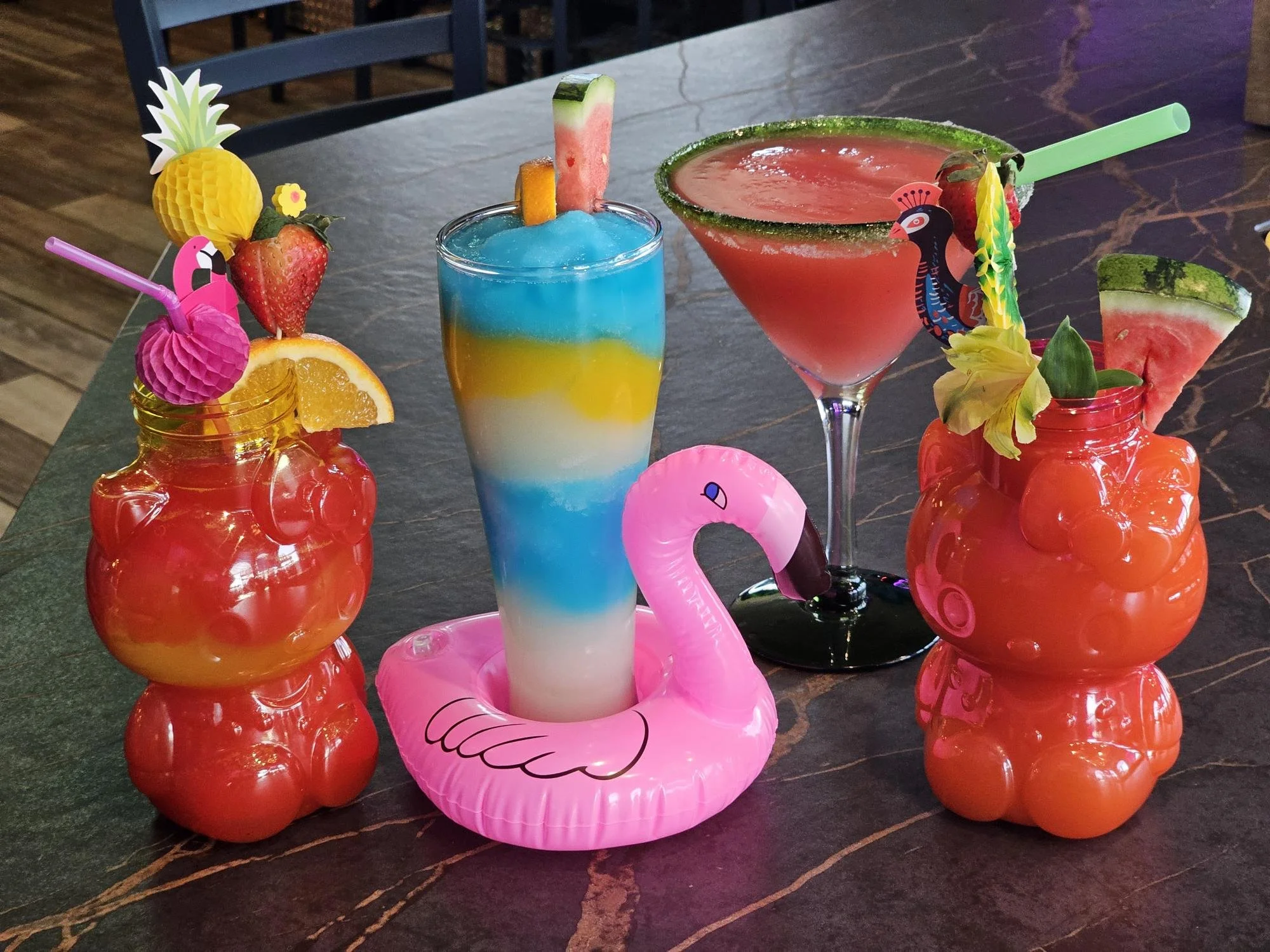 Three colorful tropical drinks with fruit garnishes and small paper decorations, set on a dark wooden table.