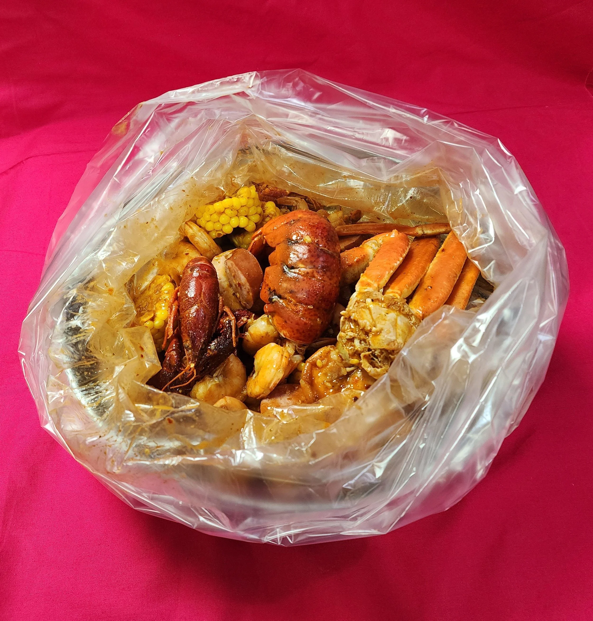Various seafood items in a clear plastic bag, including crawfish, clams, crab, and corn on the cob, placed on a pink surface.