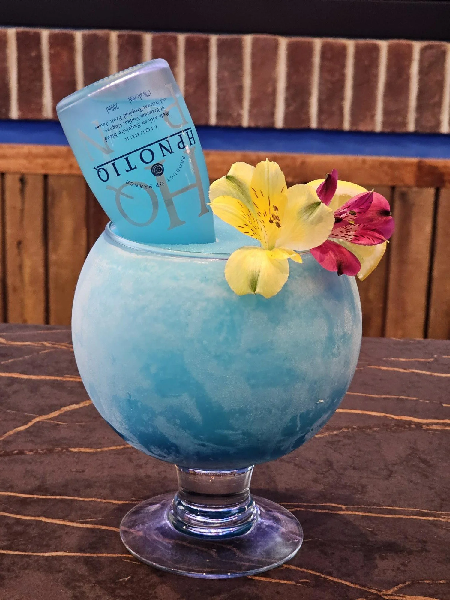 A blue cocktail garnished with yellow and pink flowers, with a small blue packet labeled 'Hydro' placed on top, in a rounded glass on a wooden surface against a brick wall.