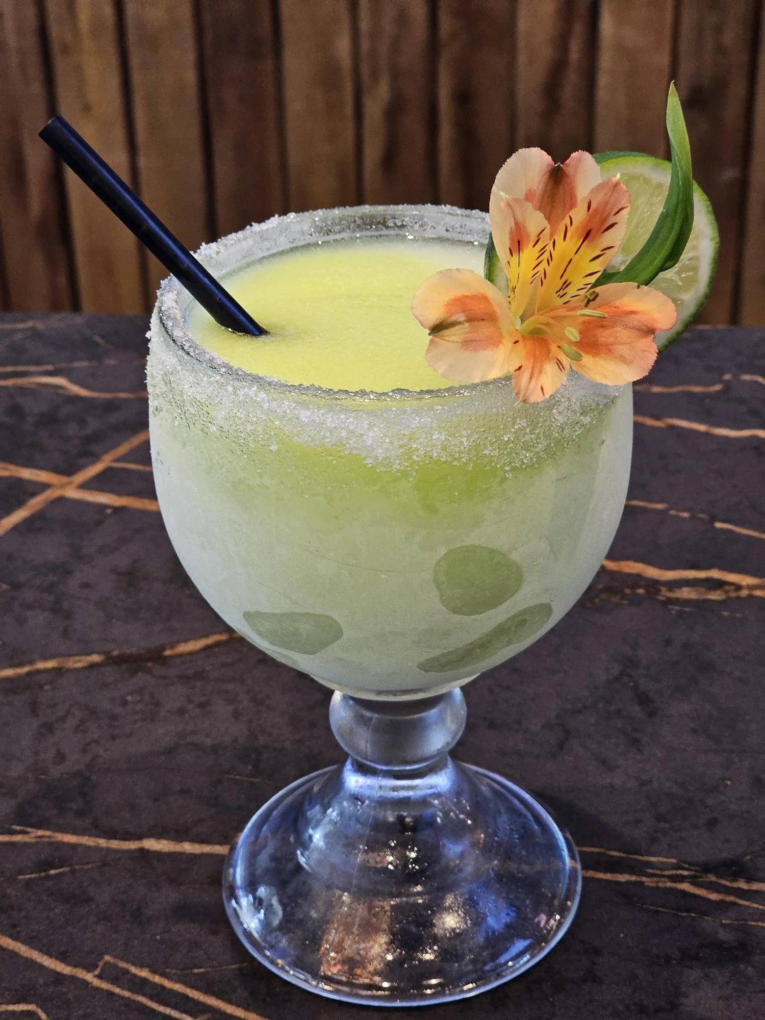 A margarita garnished with a lime wedge and an orange flower in a snifter glass, with salted rim, on a dark surface.