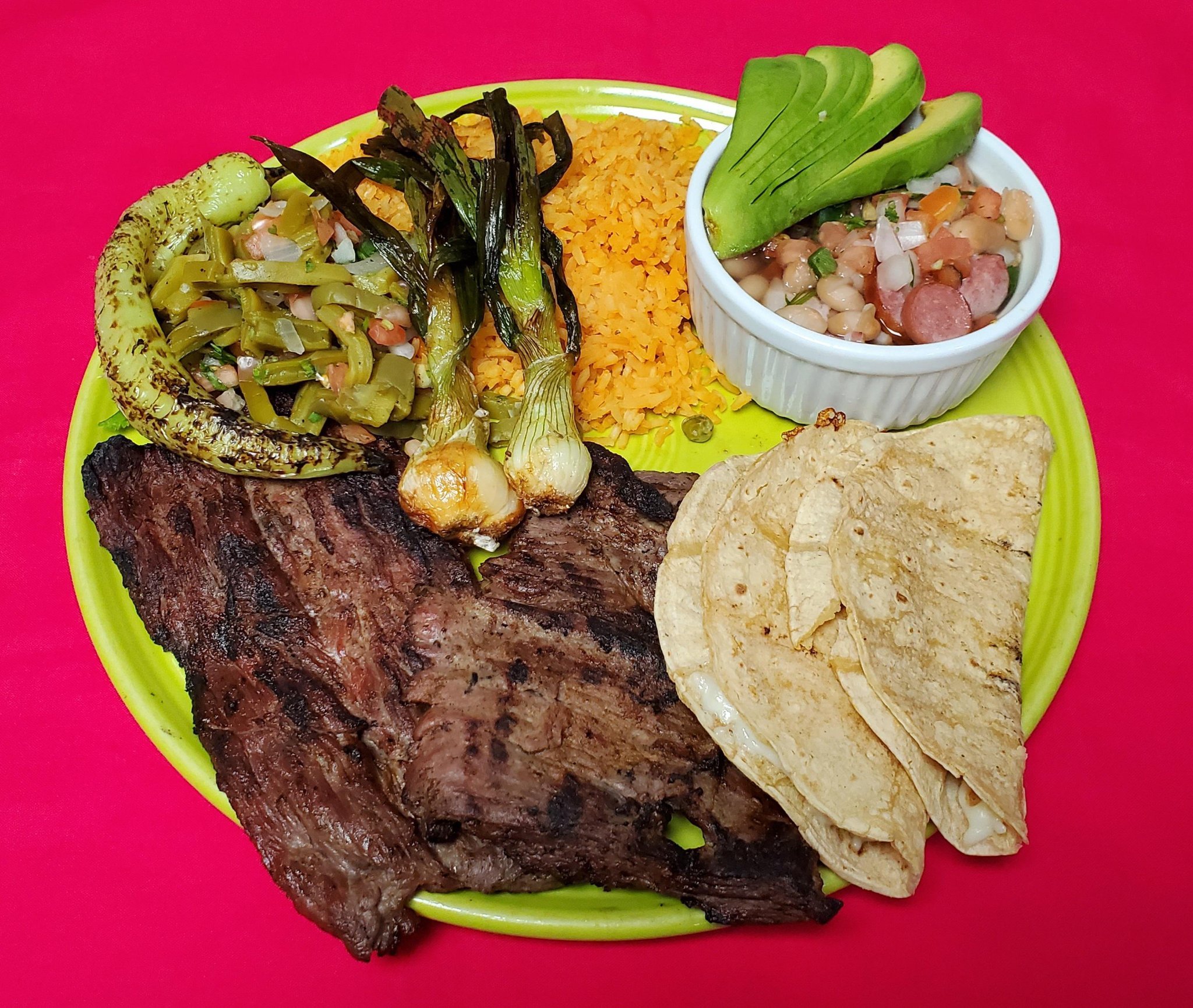 A colorful plate of Mexican food with grilled meat, tacos, roasted green onions, roasted green peppers, yellow rice, a bean and corn salad, and avocado slices on a bright pink background.