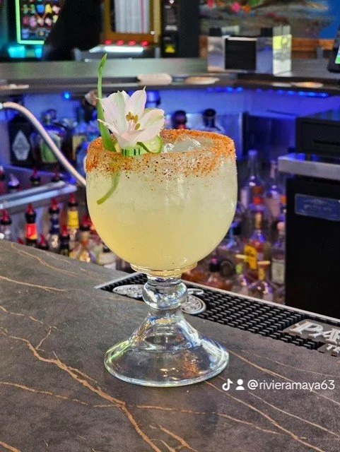 A cocktail with a salted rim, garnished with an edible flower and green leaves, on a bar counter with bottles in the background.