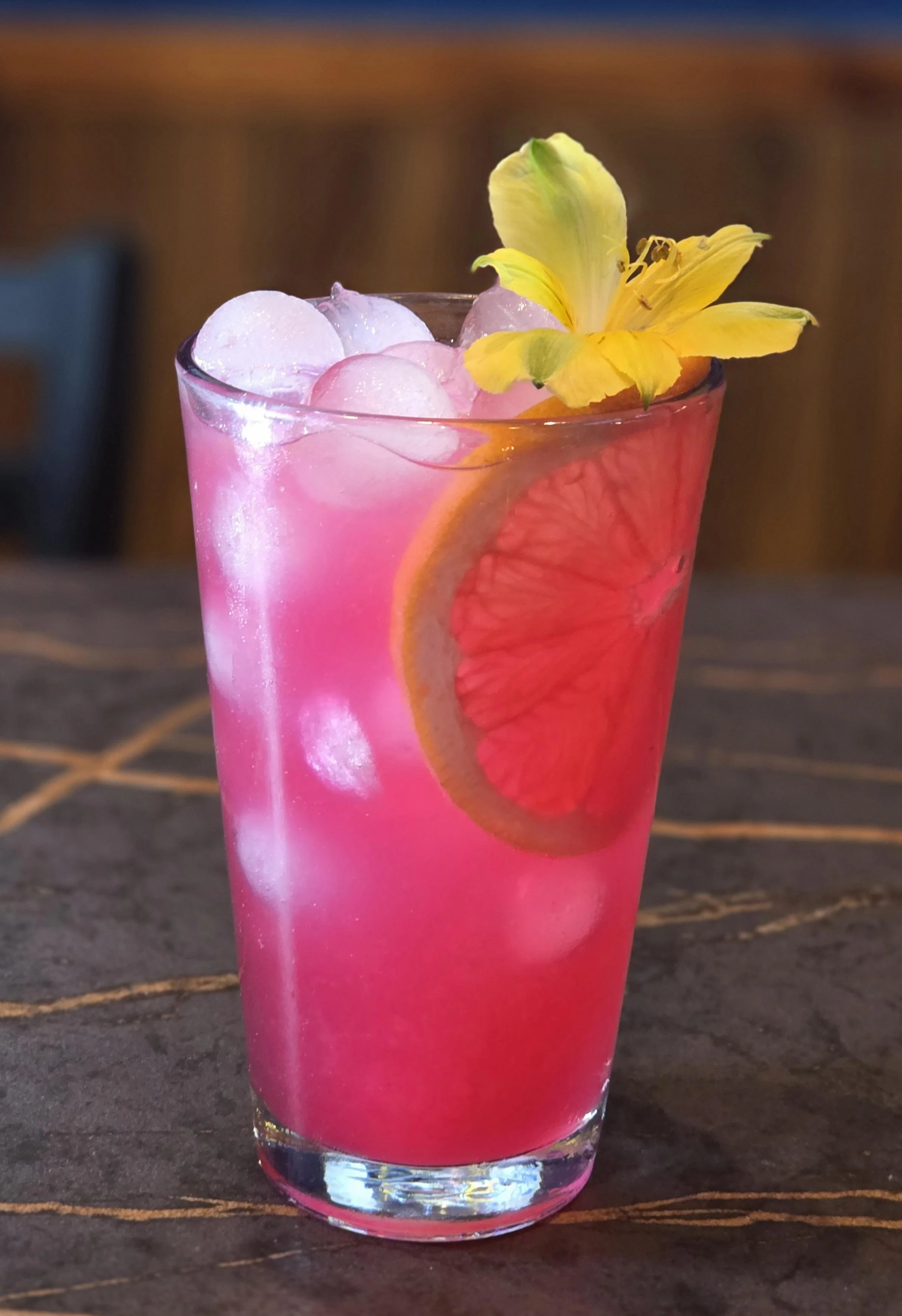 A pink grapefruit cocktail garnished with a yellow flower, a slice of grapefruit, and ice cubes in a tall glass, placed on a dark wooden surface.