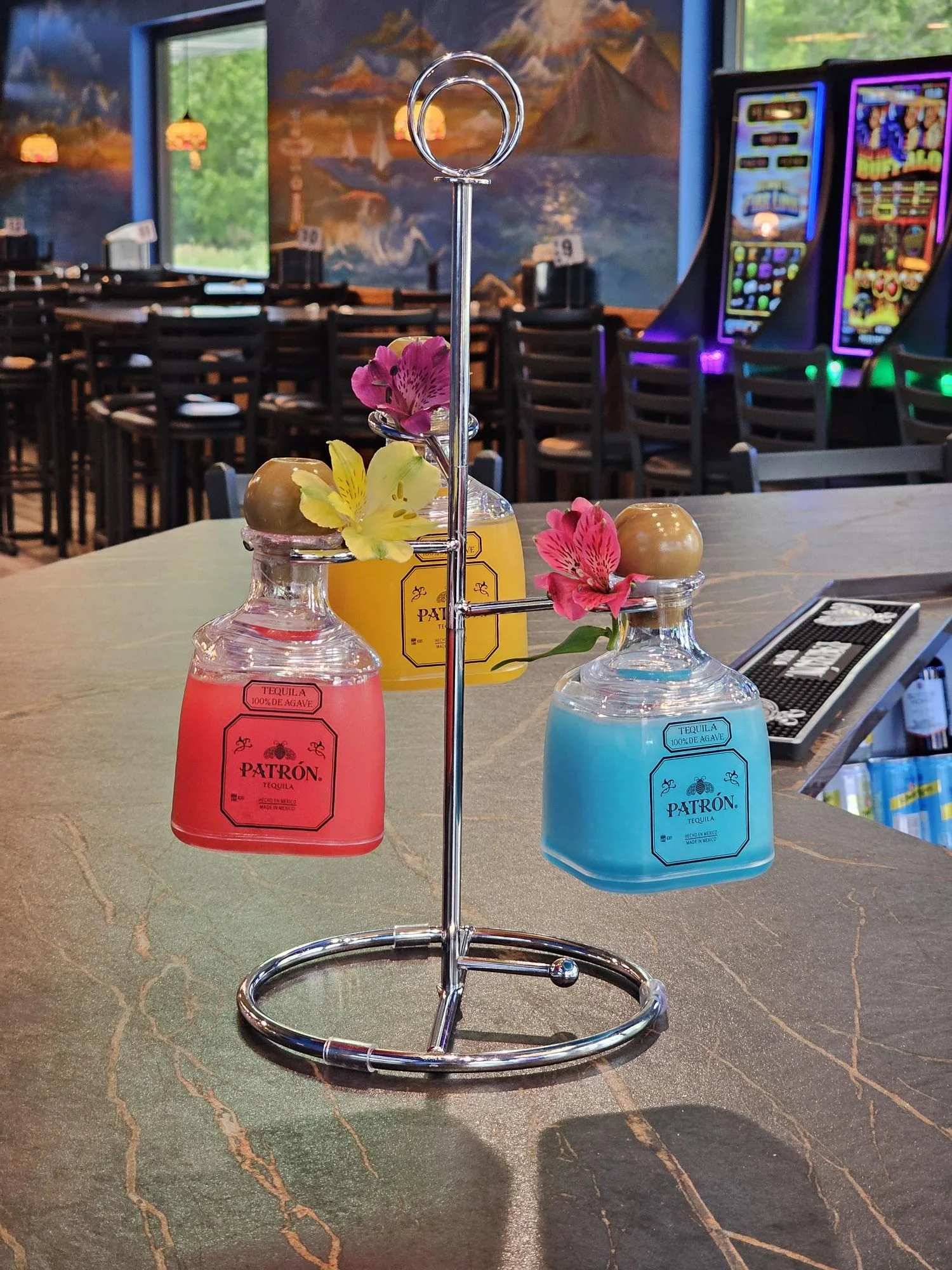 Colorful tequila bottles with flowers on top, set on a bar counter in a bar or restaurant with gaming machines and a mural in the background.