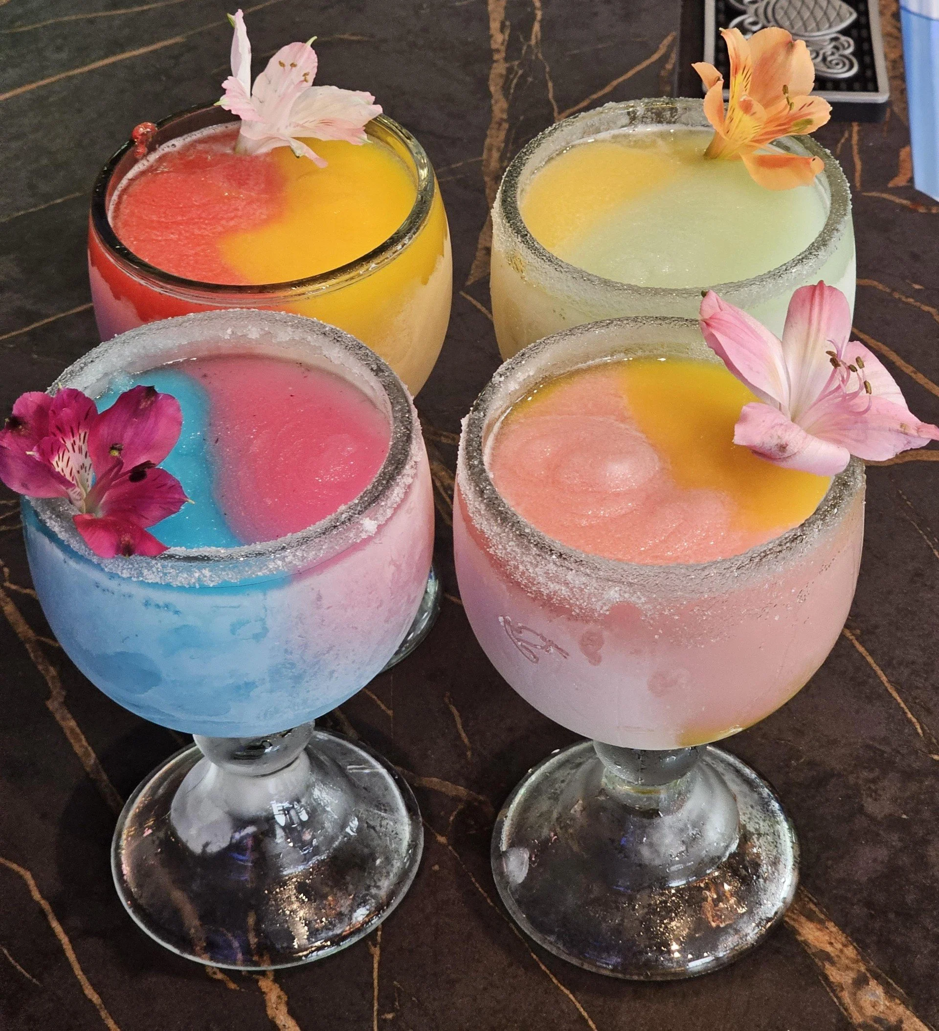 Four colorful cocktails with salted rims and edible flowers, served in large stemmed glasses on a dark table.