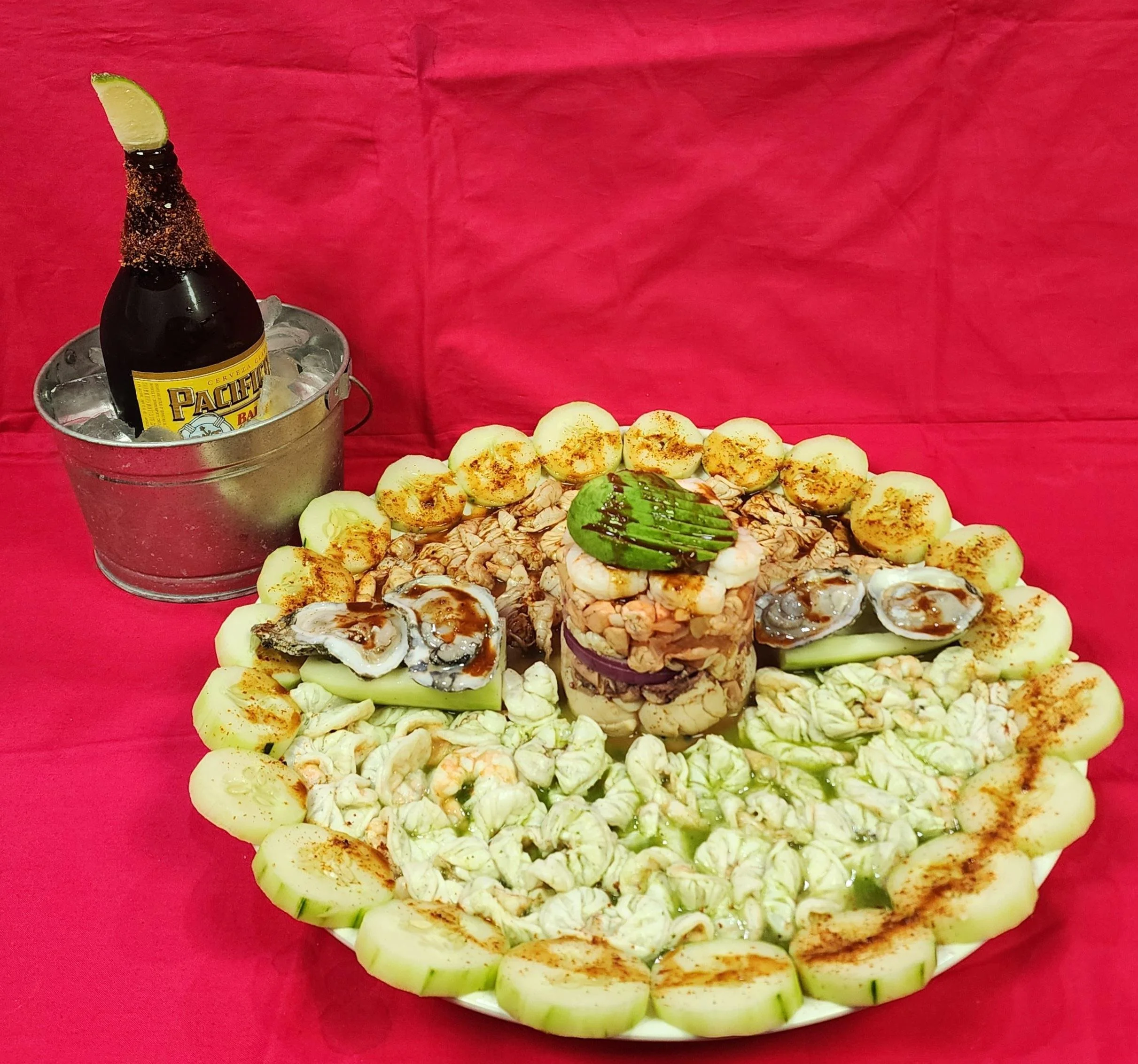 Mexican seafood cocktail on a decorative platter with sliced cucumber, lemon wedges, oysters, shrimp, ceviche, avocado slices, and a lime on top, served with a bottle of beer in a bucket of ice.