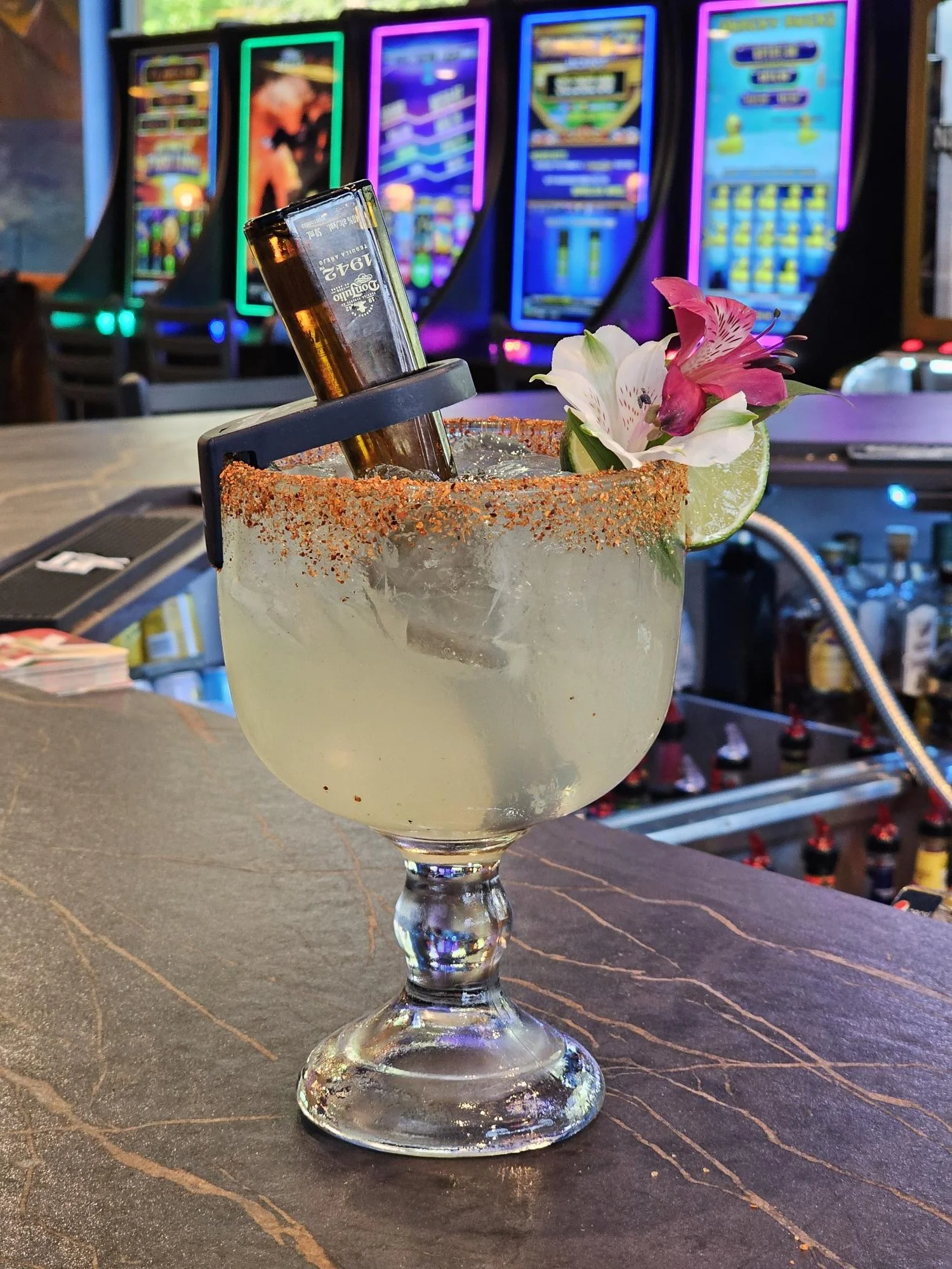 A margarita cocktail in a large glass garnished with a lime wedge and pink flower, with a salt and chili rim, on a bar counter with slot machines in the background.