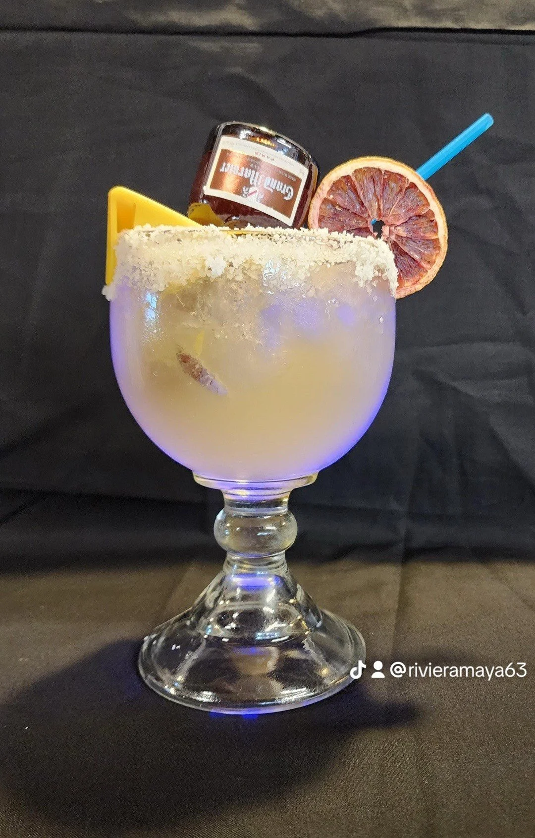 A cocktail in a large glass garnished with a lemon wedge, a dried blood orange slice, and a tamarind candy, with a small jar of cinnamon jam and a blue straw inside.
