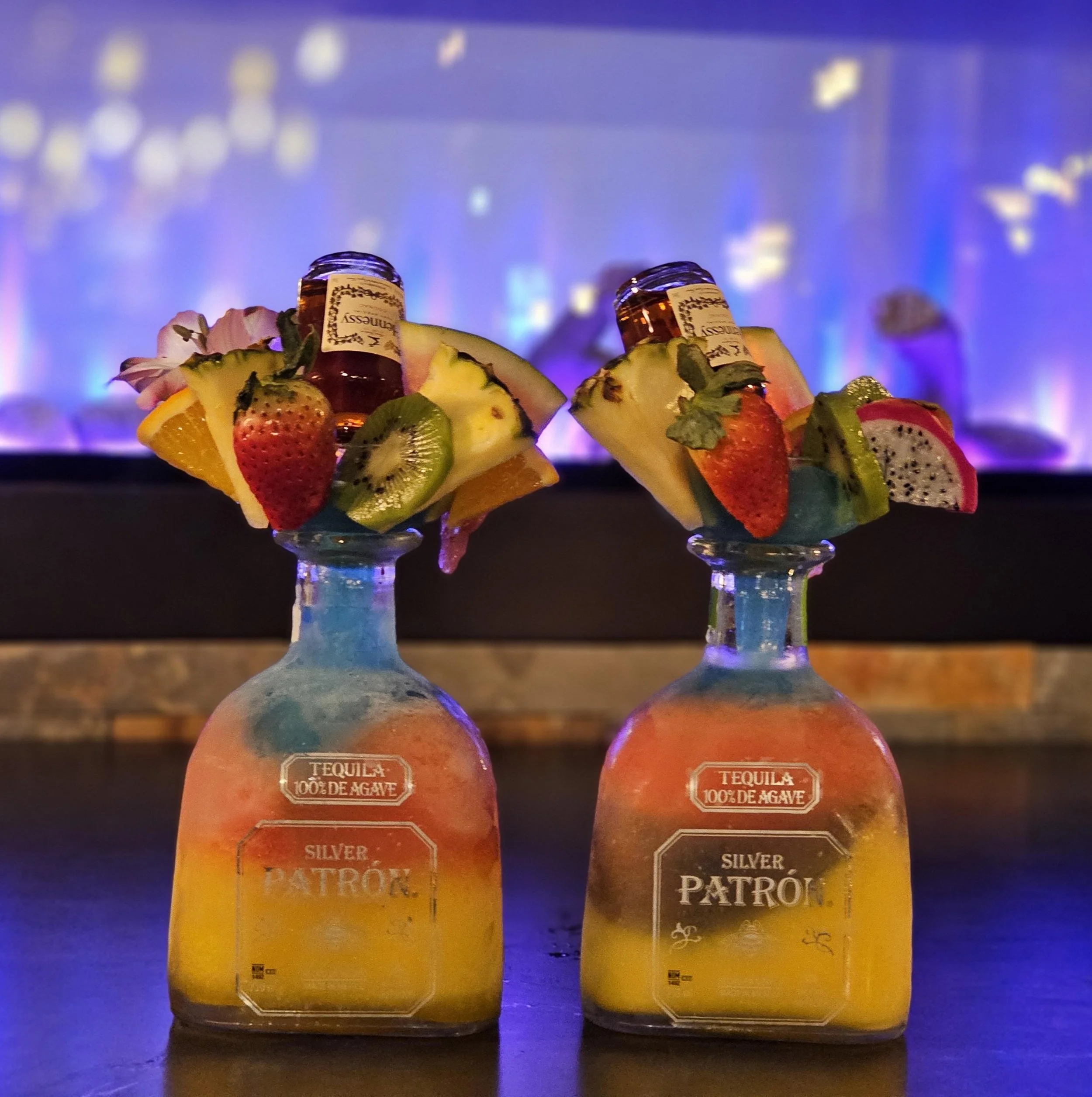 Two bottles labeled Silver Patrón tequila with tropical fruit garnishes and flower decorations on top, set against a blurred colorful background.