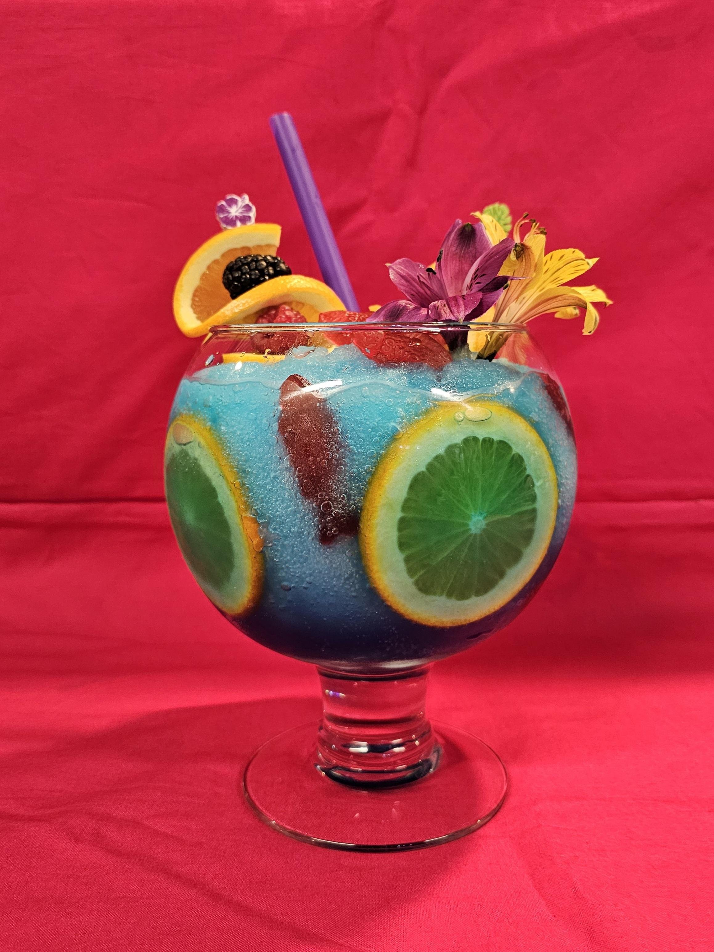 A colorful cocktail with lemon, lime, and orange slices, berries, and edible flowers in a glass with a purple straw against a bright pink background.